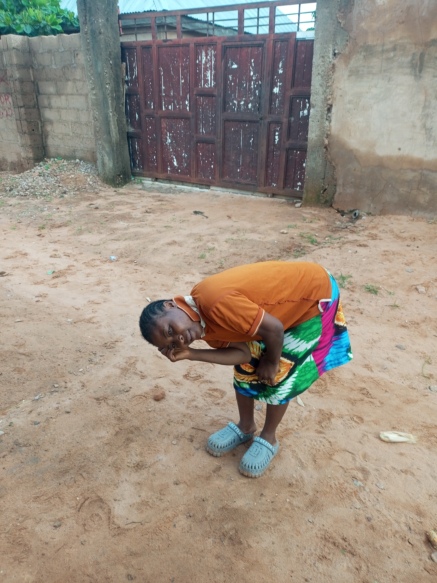 A medium shot captures a woman bending over in a sandy, outdoor area. She has dark skin, dark hair braided in cornrows, and is wearing an orange shirt with a collar and a colorful, patterned skirt. Her right hand is on her face, and she appears to be looking down. She is wearing light gray slip-on shoes with the words "GO BETTER" embossed on them. The background features a textured, reddish-brown metal gate with a white-painted grid design on top. To the left of the gate is a gray brick wall with a concrete pillar in front of it. The ground is composed of loose sand with scattered small pebbles and some dry grass. The overall lighting suggests an overcast or diffused daylight.