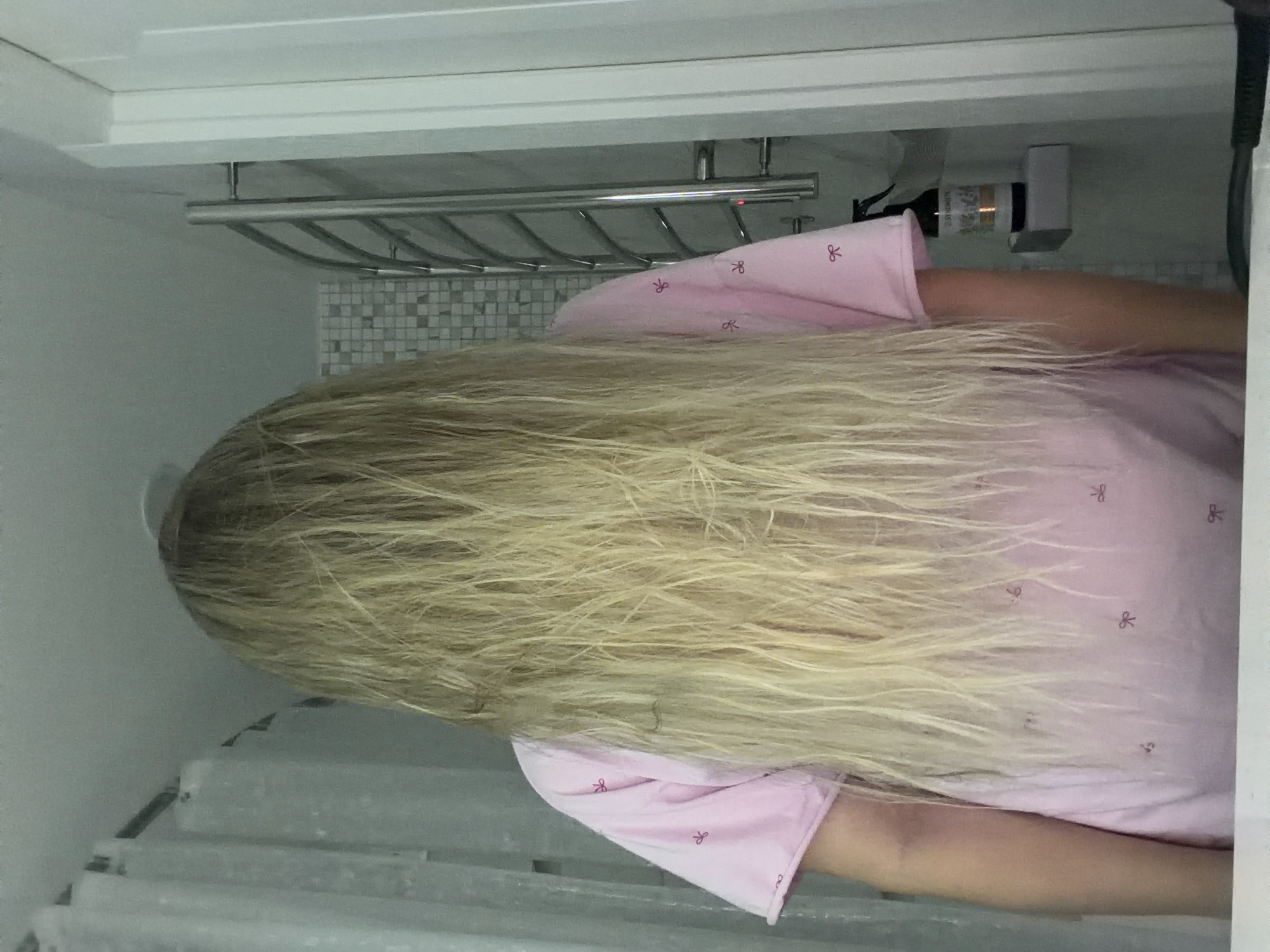 The image shows a person with long, blonde, slightly wet hair lying on a bathroom floor in what appears to be a residential setting. The person is wearing a pink shirt or pajama top with a small bow pattern. Above the person is a metal towel rack and what seems to be a mosaic tiled wall. A portion of a white shelving unit or cabinet is visible at the top of the frame, and possibly a hair care product is situated on a shelf. The lower part of the image reveals a white, slatted floor. The image doesn't provide sufficient information to pinpoint the location to Votkinsk, Russia. The forensic description details the elements visible in the photograph.