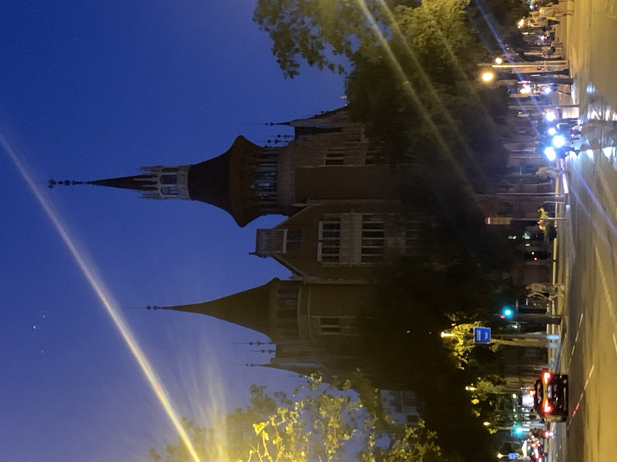 The image depicts an urban scene at dusk, with the sky a deep twilight blue. In the foreground, the distinct architectural style of a building, possibly a historic landmark, is visible. It features multiple turrets and pointed roofs, suggesting a Gothic or Modernist design, consistent with Barcelona's architecture. The building appears to be illuminated, with light emanating from its windows.
Adjacent to the building is a street with traffic. Several cars and motorcycles are present, their headlights and taillights creating streaks of light due to the long exposure or low light conditions. Some people are seen standing on the sidewalk and crossing the street, appearing as silhouettes against the illuminated background. Trees with sparse foliage line the street, their leaves catching the ambient light.
Notable details include the overall low light setting, indicative of evening or early morning. The presence of streetlights and illuminated buildings confirms the urban environment. The architectural style of the building strongly suggests a location in Barcelona, Spain. No visible text is discernable in the image. The scene appears to be a bustling city street at a quiet time of day, perhaps after sunset, with a mix of architectural grandeur and everyday urban life.