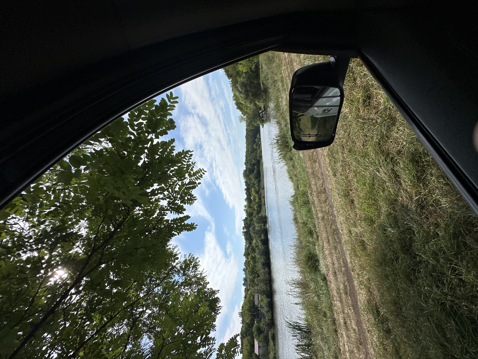 The image depicts a view from inside a car, looking out towards a tranquil body of water. The foreground is dominated by the interior of the car, including the car door and a side-view mirror reflecting a white vehicle. Beyond the car, a grassy embankment slopes down to a wide river. The river is calm, with gentle ripples on its surface, and is bordered by lush green trees on the opposite bank, creating a dense, natural landscape. Several scattered houses with red roofs are visible nestled among the trees on the far side of the river, suggesting a rural or semi-rural setting. The sky is partly cloudy, with fluffy white clouds scattered across a bright blue expanse. Sunlight filters through the leaves of a large tree in the upper left corner of the image, casting dappled shadows. The scene appears to be set in Makiv, Ukraine, given the context provided.