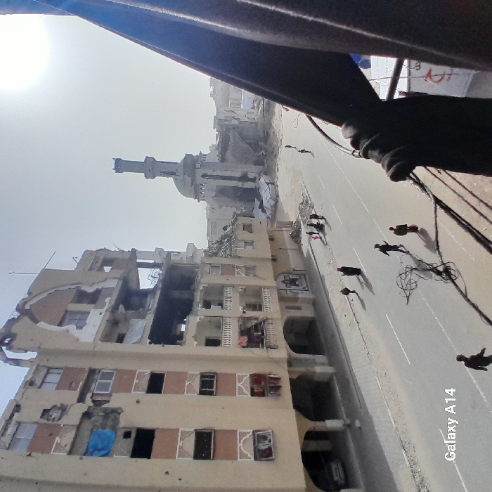 A war-torn street scene with buildings extensively damaged, some reduced to rubble. A mosque with a minaret stands amidst the destruction. Several people walk along the road, casting shadows. The image is taken from an elevated perspective, possibly from inside a building, with part of the window frame visible. The light is harsh, and the sky is hazy. A "Galaxy A14" watermark is present at the bottom right corner.