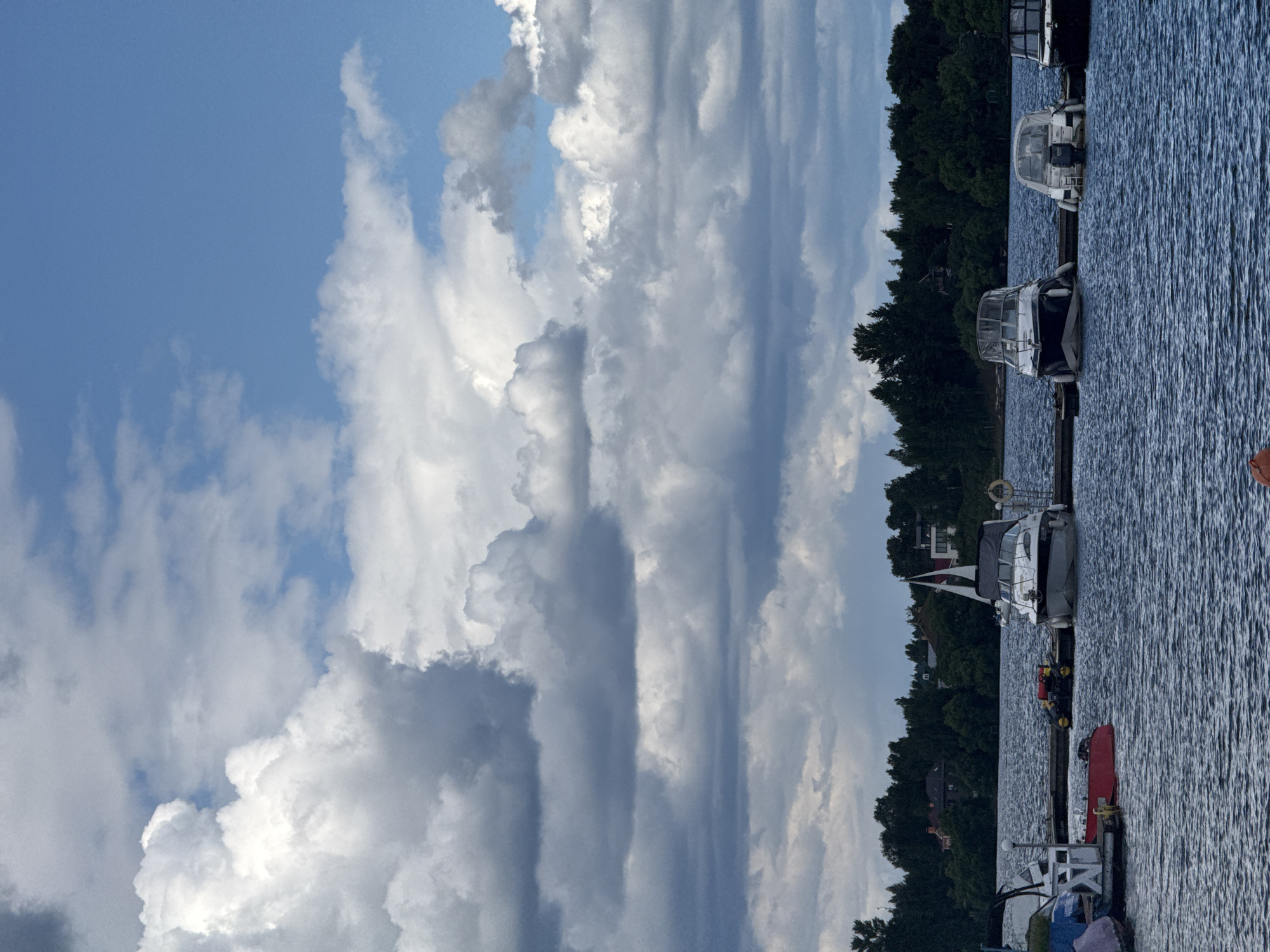 A panoramic, eye-level shot captures a serene waterscape at the Tourist Boarding House "Klyazminskoye Reservoir," Russia. The scene is dominated by a vast expanse of deep blue water, reflecting the dramatic, cloud-filled sky above. A line of boats is docked along a wooden pier in the foreground, their hulls varied in color and size. The largest boat, a white motor yacht with a black canopy, is positioned centrally, its sail furled. To its left, a smaller white boat with a red hull and a white structure is moored. Further to the right, two other motorboats, one with a white canopy and another with a dark hull, are tied up. The background features a dense line of dark green trees, behind which several houses with red roofs are partially visible. The sky is a dynamic mix of bright blue patches and large, fluffy white and gray clouds, suggesting a day with changing weather. The overall impression is one of a peaceful, natural setting on a body of water, with the docked boats indicating a recreational or residential area.