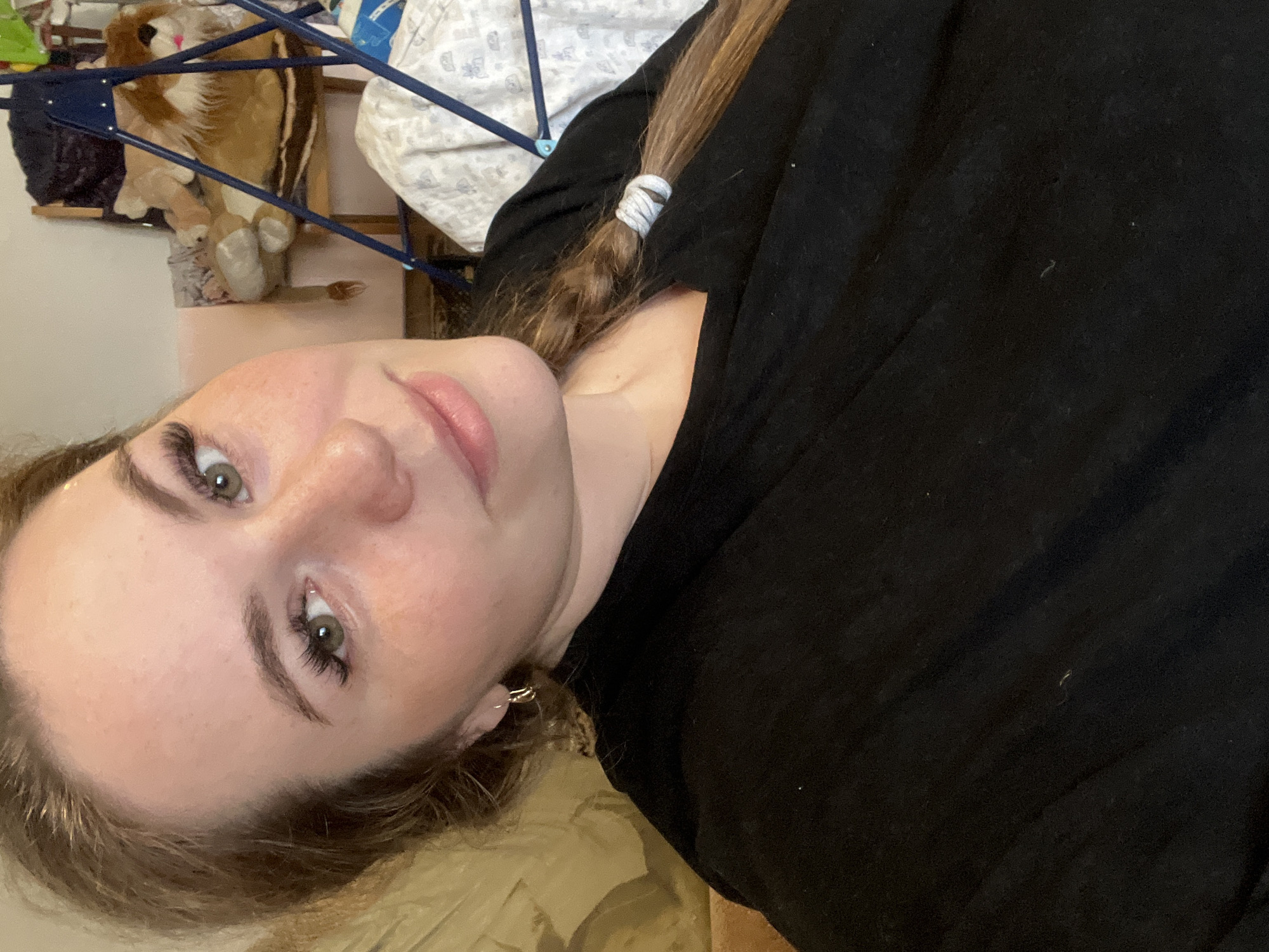 A selfie portrait of a fair-skinned person with light brown hair styled in a braid, wearing a black shirt. The background includes a stuffed lion toy, laundry rack, and bedding. The location in the city of Сосново, Russia, cannot be determined from the image.