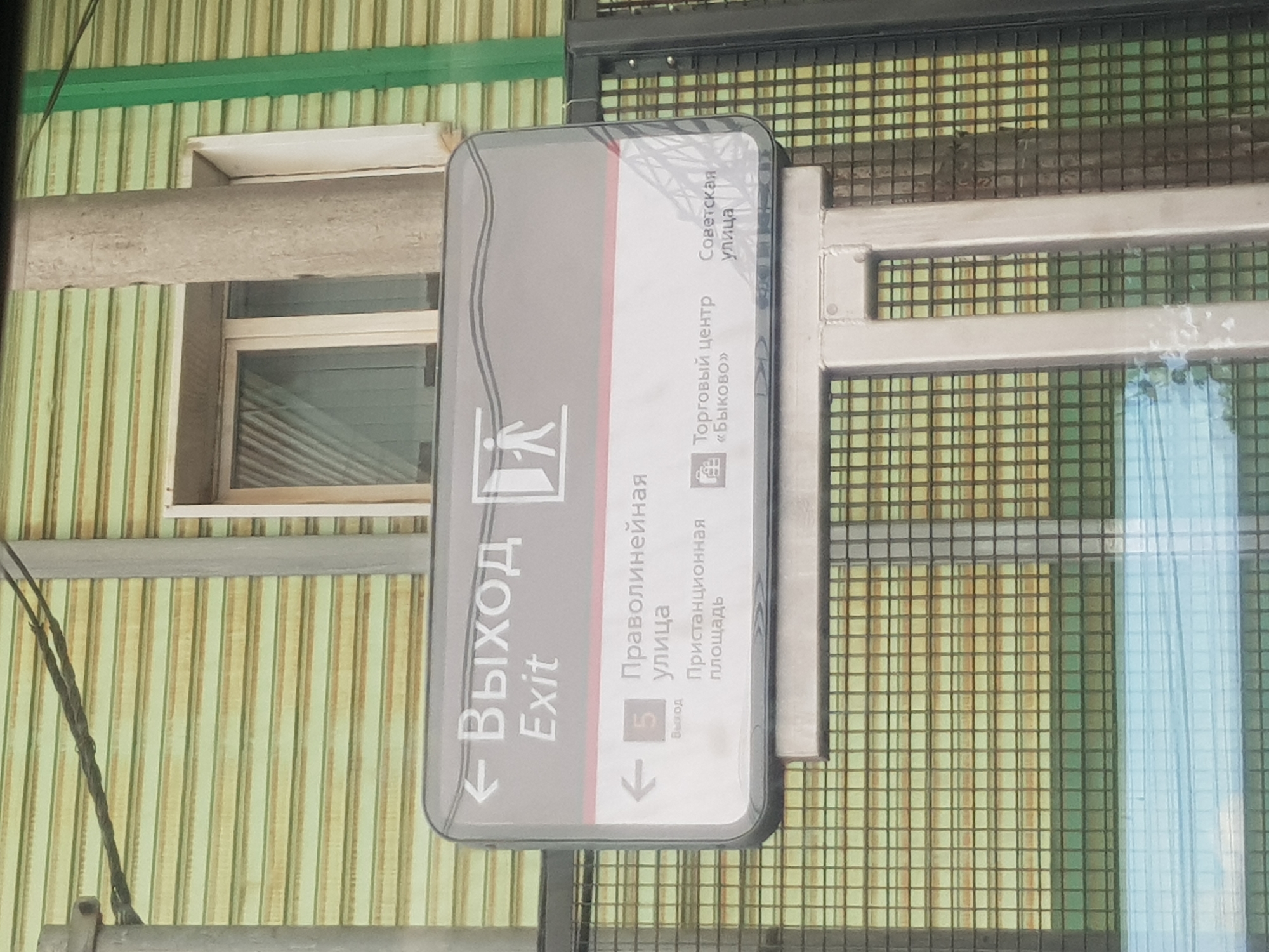 Here is a detailed description of the image:
**Scene Description:**
The image depicts an exit sign written in both Russian and English, indicating directions to several locations. The sign is mounted on a metal framework with a wire mesh background.
**Sign Details:**
* **Top Line:** "Выход" (Russian for Exit) and "Exit" (English). An arrow points to the left, indicating the direction.
* **Second Section:** A symbol of a walking person, indicating the direction of exit.
* **Bottom Section:** Lists directions: "Праволинейная улица" (Pravolinieynaya Street), "Пристанционная площадь" (Pristantsionnaya Square), "Торговый центр «Быково»" (Bykovo Shopping Center) and "Советская улица" (Sovetskaya Street), along with an exit number 5.
**Background:**
The background behind the sign consists of a corrugated metal wall with faded vertical stripes in light green and yellow. There is a window with a white frame and a concrete slab-like structure above it. The metal framework of the sign structure is also visible.
**Overall Impression:**
The sign appears to be a directional sign indicating the way to exit and directing people to various locations within the vicinity. The general appearance suggests a location within Russia, based on the presence of Russian text.