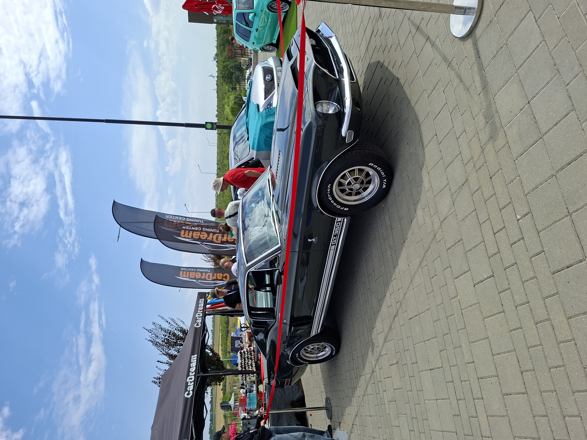 The image depicts an outdoor event, likely a car show, on a sunny day. In the foreground, a dark-colored vintage car, identified as a GT. 350 by its badging, is prominently displayed. The car is sectioned off by a red ribbon, held by stanchions. Several individuals are visible, some appearing to be attendees and others possibly staff. To the left, a black canopy tent with "CarDream" branding is set up, and further back, tall, dark banners with "CarDream TUNING CENTER" are erected. In the mid-ground, other vehicles are visible, including a silver sedan covered with a turquoise cloth and a bright teal hatchback. The ground is paved with light-colored interlocking bricks, and the background shows a line of trees and a clear sky with scattered clouds. The setting suggests a public gathering, possibly in a parking lot or open area, consistent with the rural surroundings of Stăuceni, Moldova.
