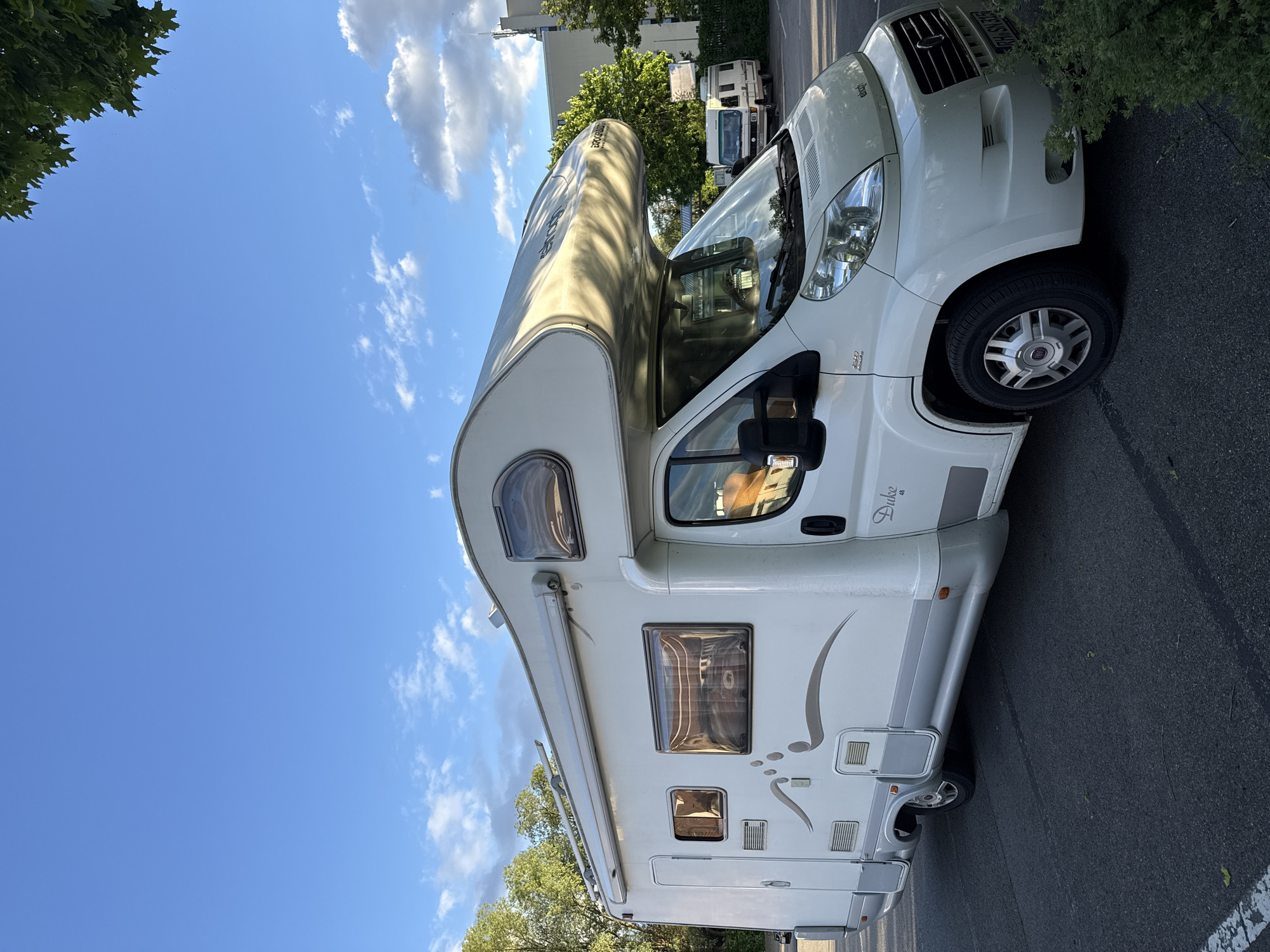 The image depicts a white recreational vehicle (RV) parked on an asphalt road in Riedlingen, Germany. The RV is a "Duke" model. The sky is blue with scattered clouds. Green trees are visible in the background, along with a building and another RV in the distance. The street is a parking area of the city of Riedlingen. There are no visible people.