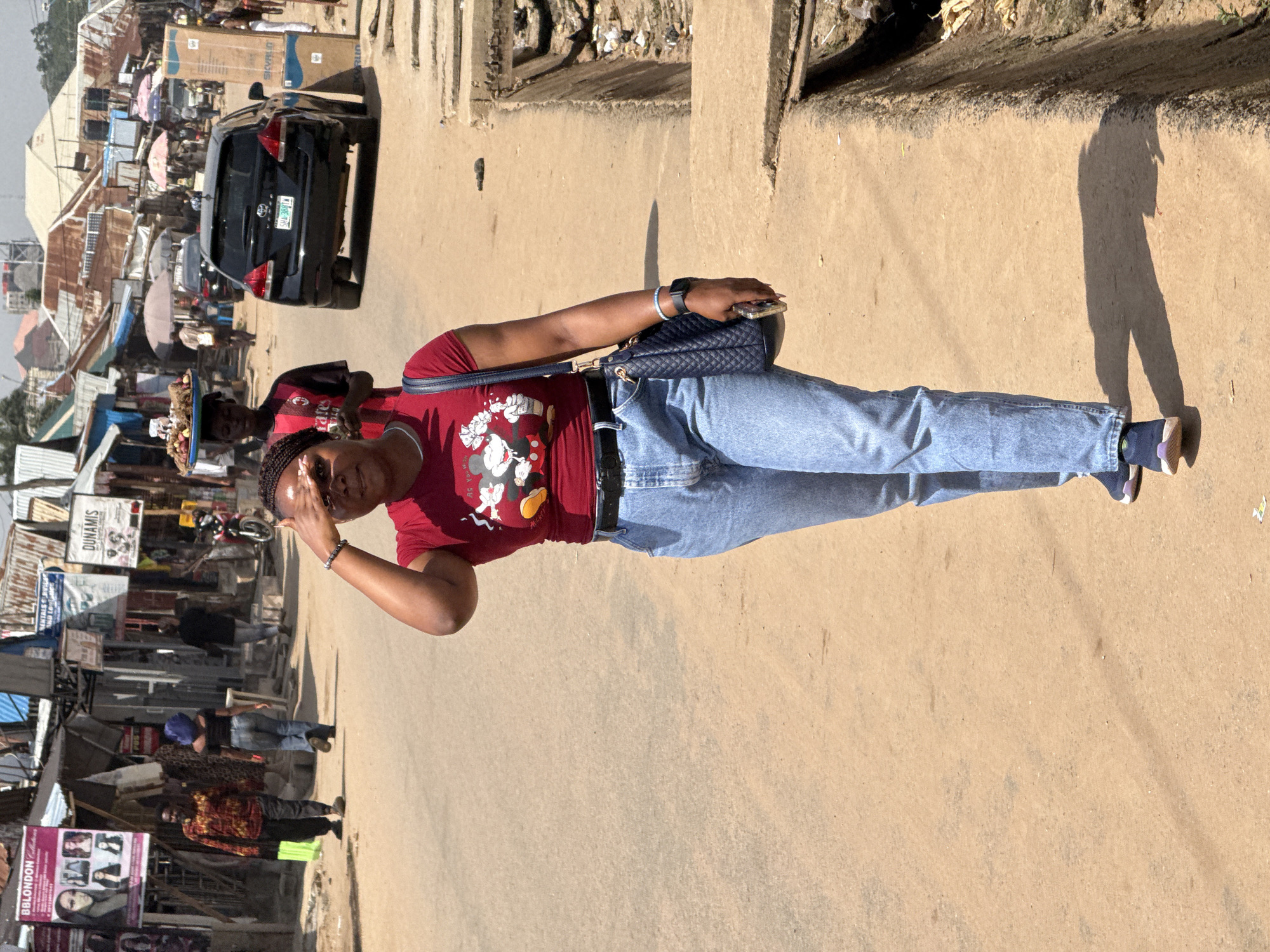 A woman is walking on a dirt road in an open-air market in Abuja, Nigeria. She is wearing a red t-shirt with a Mickey Mouse graphic, blue jeans, and dark sneakers. She is carrying a dark blue purse on her right shoulder and holding her left hand up to shield her eyes from the sun. In the background, there are various market stalls with corrugated metal roofs, signs, and merchandise displayed. A black SUV is parked on the road, and several other people are visible walking in the distance, including a person carrying a tray of food on their head. The ground is dusty, and a gutter runs along the side of the road.