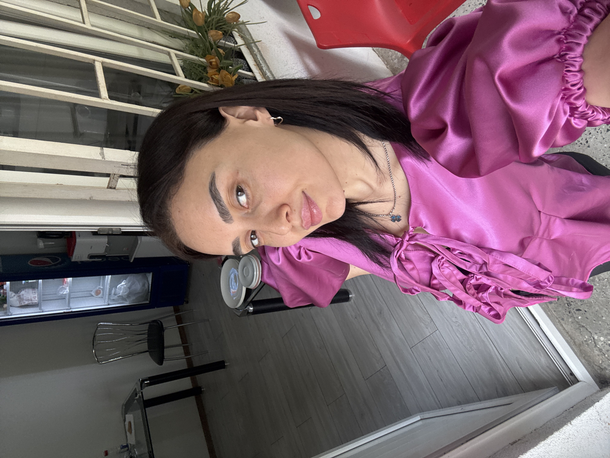 A woman with dark hair, wearing a purple satin blouse and a necklace, is posing for a photo. She has light skin, dark eyebrows, and is wearing earrings. The background shows a room with grey wooden floors, a table and chair, and a blue Pepsi cooler with drinks inside. Outside the window, there is a window with plants on the windowsill and a red chair. The location is in Tbilisi, Georgia, based on the characteristics of the environment.