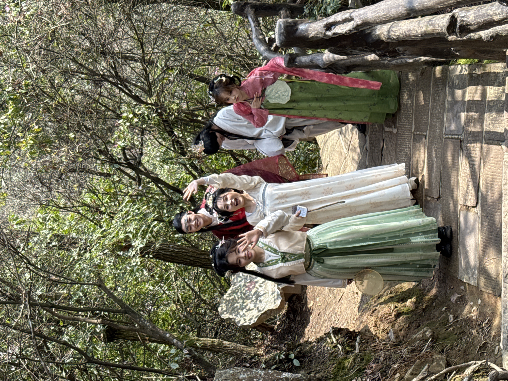 Five individuals, comprising three females and two males, are depicted on a stone-slab path within a densely wooded area of Wulingyuan, China. The scene is illuminated by direct daylight, casting shadows across the path.
In the foreground and mid-ground, three individuals face the camera, smiling and gesturing with raised hands. From left to right:
1. A female in a light green and off-white traditional outfit, with dark hair styled with side buns, waves her right hand. She holds a round, light-colored fan in her left hand.
2. A female in a cream-colored traditional dress, with floral hair decorations, waves her right hand high above her head. She holds a small, light-colored rectangular object in her left hand.
3. A male in a dark red robe with a dark headband, featuring a light-colored band across his forehead, waves his right hand.
To the right of the path, a rustic wooden railing, constructed from natural or carved logs, is partially visible. A female in a pink top over a green long skirt leans against this railing, holding a large, leaf-shaped fan against her chest with both hands. She looks towards the camera with a slightly pouting expression. Behind her and to the right, a male in a white traditional robe, with long dark hair and a golden headpiece, stands looking downwards and to the right.
The path is composed of large, irregular stone steps. To the left, an earthen bank rises, featuring exposed roots and a prominent light-colored rock with faint, irregular markings. The background consists of dense tree foliage and numerous branches, indicating a natural, forested environment.