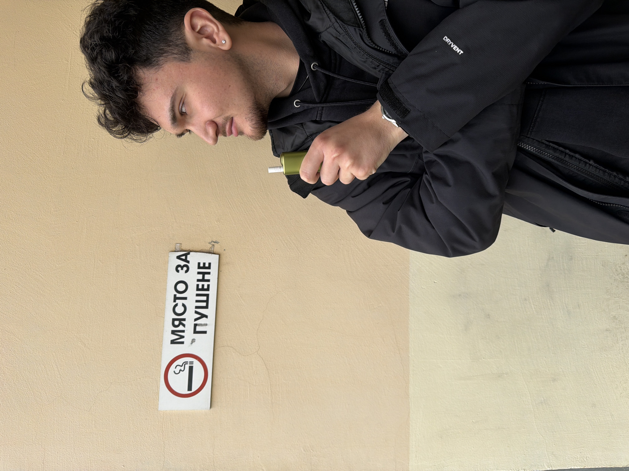 The image shows a man standing next to a wall with a smoking area sign. The man has dark curly hair and is wearing a black hooded jacket. He is holding a green object in his right hand with a white object protruding from it. The wall is two-toned, beige on the left and a lighter shade on the right. The sign on the wall reads "МЯСТО ЗА ПУШЕНЕ" in Cyrillic, which translates to "Smoking Area." The image appears to be taken in Sofia, Bulgaria, based on the language of the sign.