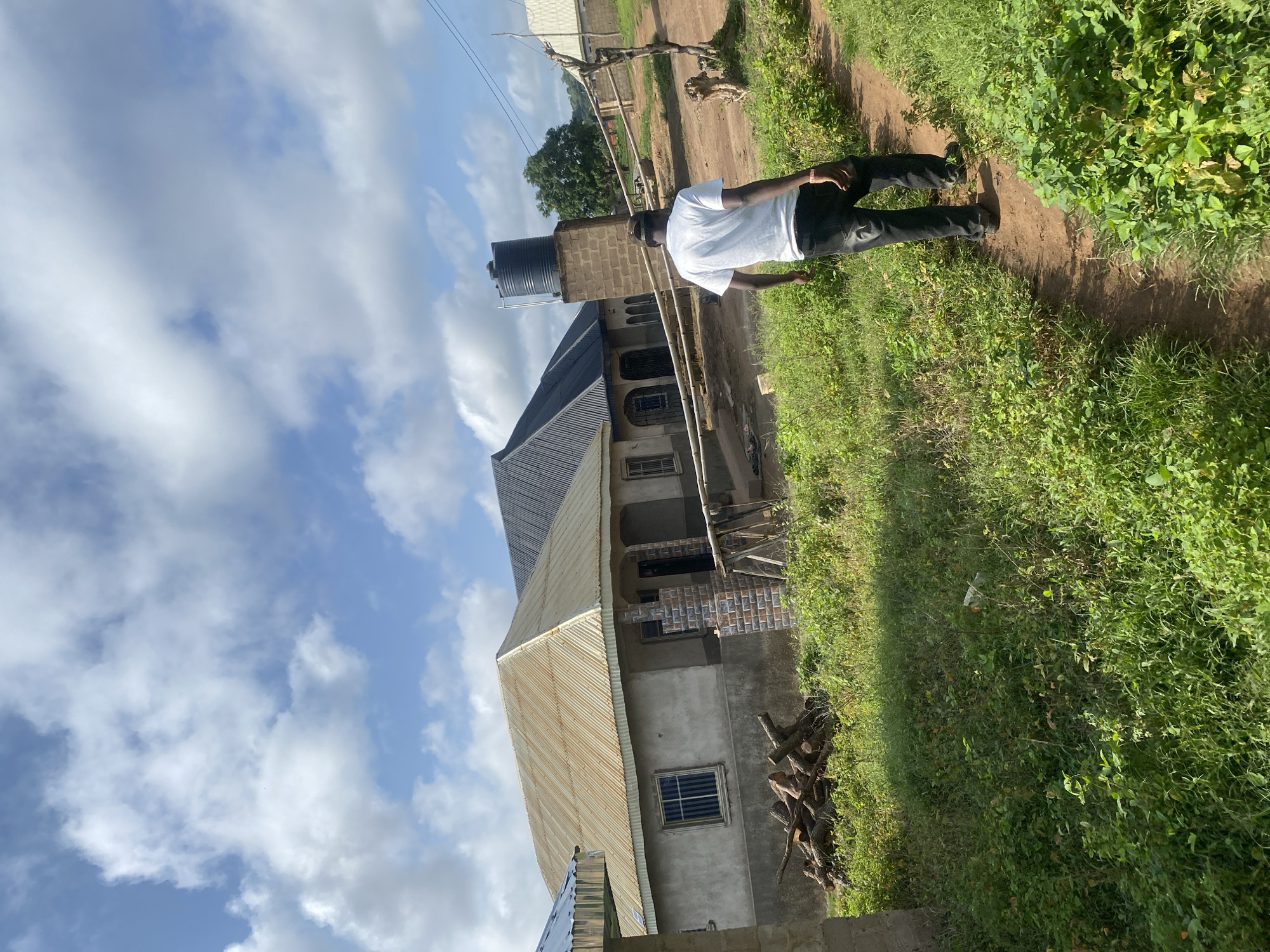 A person is standing on a dirt path in an outdoor setting. They are wearing a white t-shirt and dark pants. The person is facing away from the camera, with their right arm extended slightly. The path is bordered by green grass and some vegetation. In the background, there is a building with a corrugated metal roof and a water tank on top of a brick structure. The sky is blue with white clouds. The scene appears to be during the daytime, with clear weather. No visible text is present in the image.