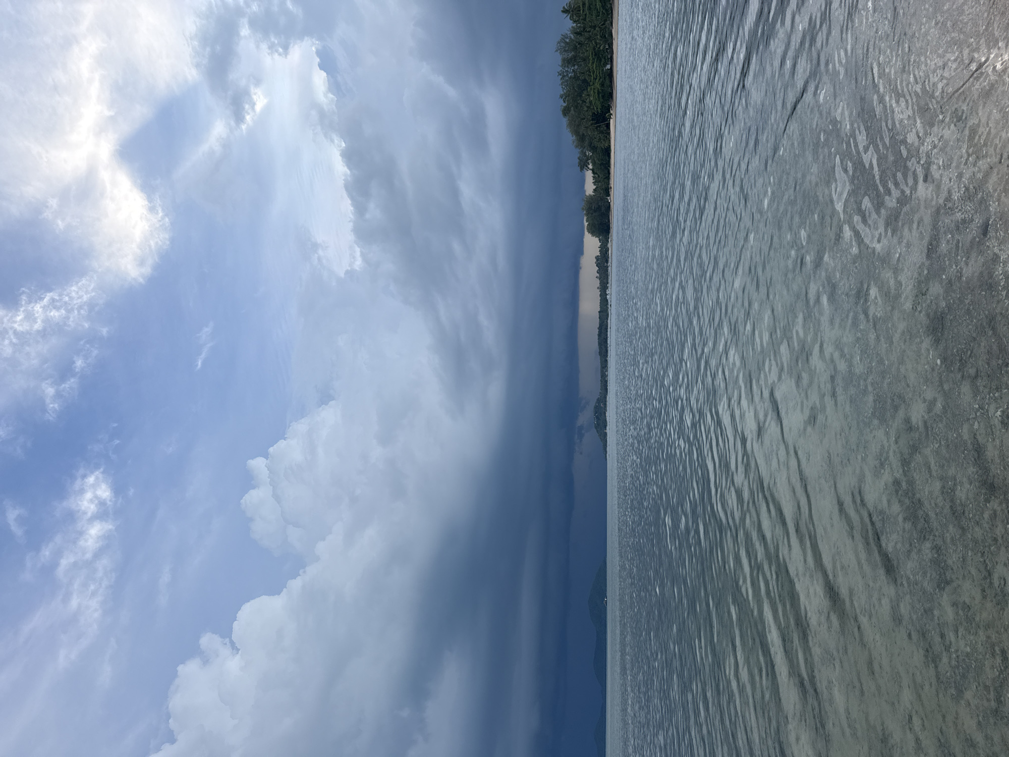 A wide shot shows a body of water with a sandy shore on the right and a treeline in the distance, beneath a dramatic sky. The water is calm, with subtle ripples breaking the surface and showing the sandy bottom beneath. Dark, heavy clouds dominate the sky, with shafts of sunlight breaking through on the upper right, illuminating patches of lighter blue sky. The treeline on the right is dense with green foliage, meeting a sandy beach. In the distance, a few low-lying hills or islands are visible, shrouded in haze and the dark clouds. No people or objects are identifiable in the image. The scene is likely located in Ban Bang Khao, Thailand, given the tropical vegetation and coastal environment.