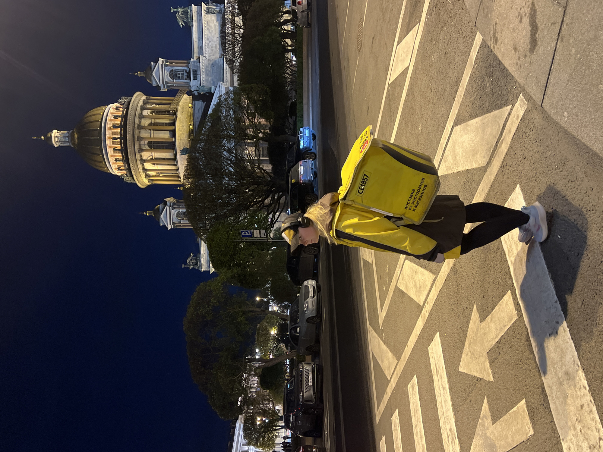 The image shows a delivery person in a yellow uniform and backpack standing on a street in Saint Petersburg, Russia. The delivery person is standing in a crosswalk with painted arrows. Behind the person, there is a row of parked cars and a street with trees on either side. In the background on the left is Saint Isaac's Cathedral. The sky is dark, suggesting it is nighttime.