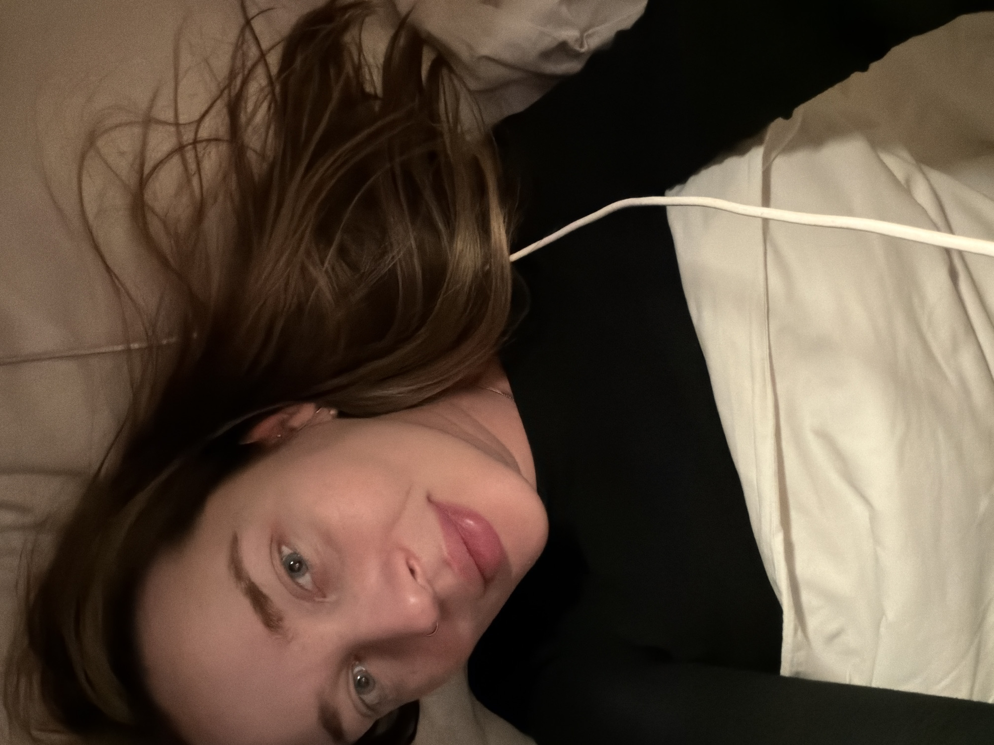 A woman with long brown hair is lying down, looking directly at the camera. Her hair is spread out on what appears to be a bed or pillow. She is wearing a black top. There is a white object, possibly a cable, running across the scene. The background consists of the bed linens and wall. The setting does not show Moscow, Russia.
