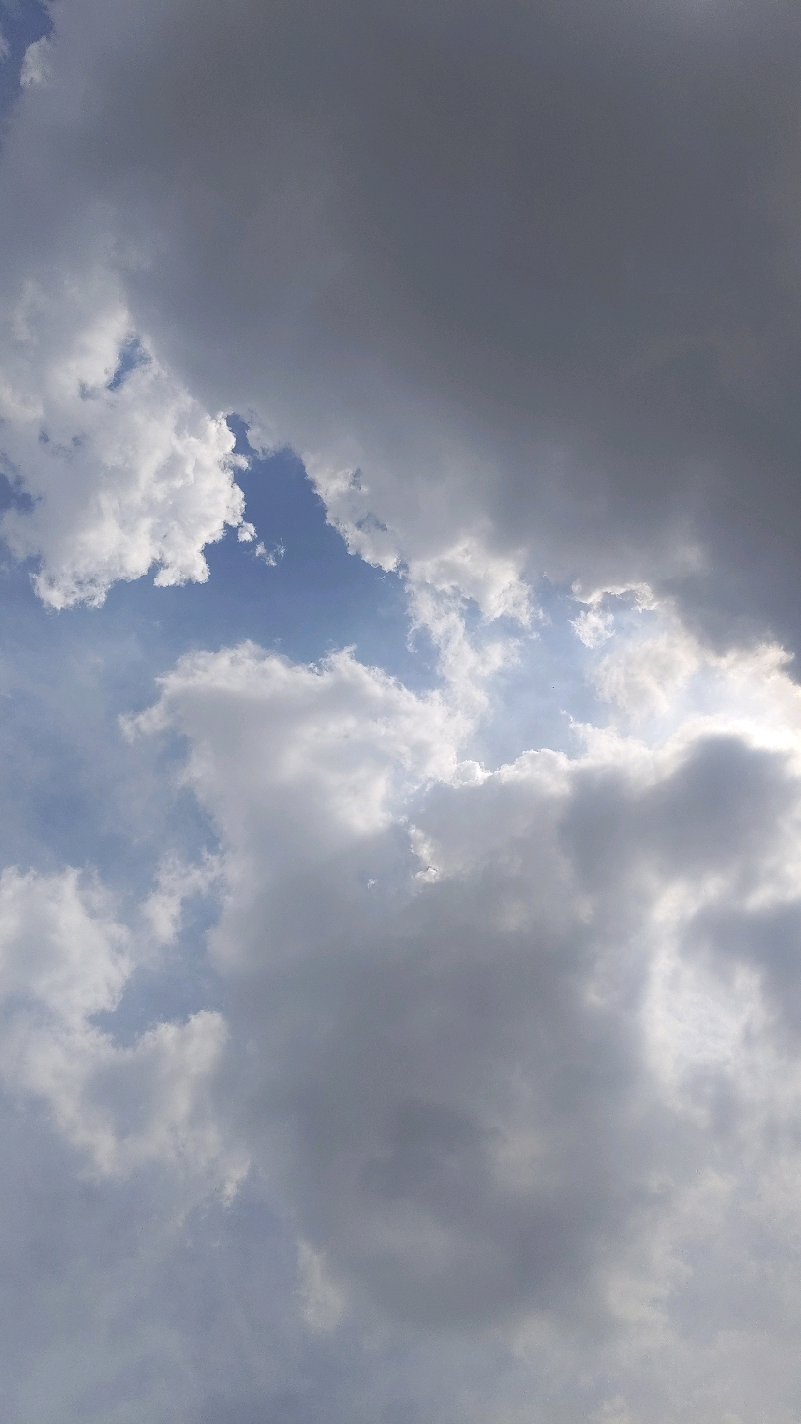 The image is an outdoor, vertical shot of a cloudy sky with patches of blue visible. The clouds vary in density and color, ranging from bright white to dark gray, suggesting a mix of cumulus and possibly nimbostratus formations. Sunlight is breaking through in some areas, illuminating the edges of the clouds and creating bright highlights. The overall scene depicts a dynamic sky, likely captured during daylight hours. There are no people, animals, structures, or discernible landmarks visible in the image.