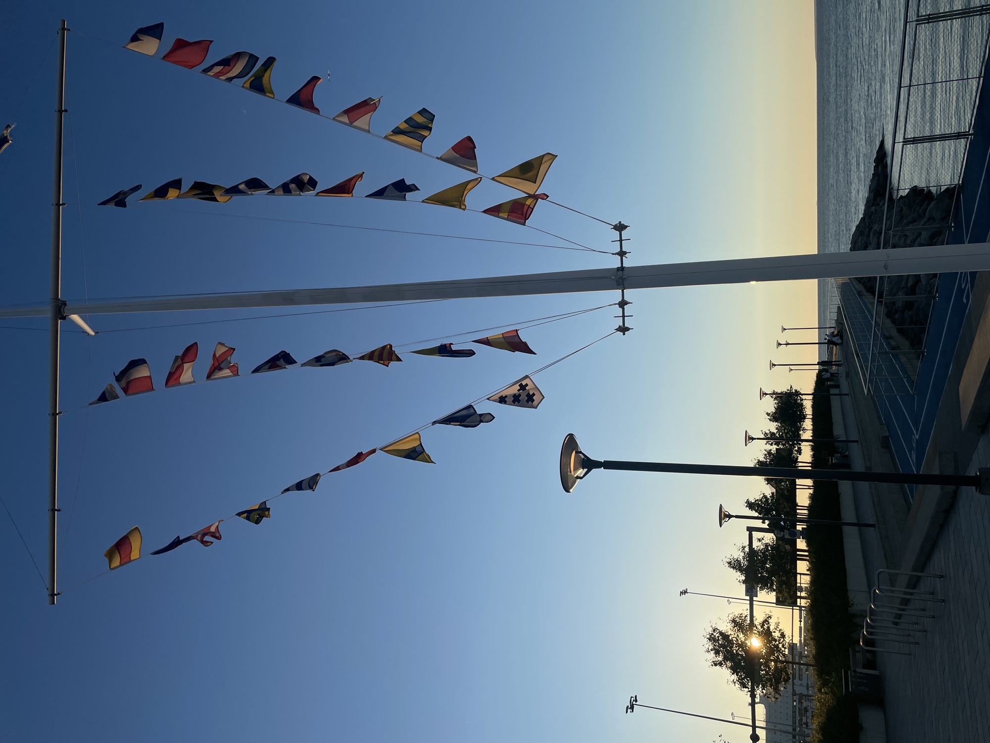An outdoor scene at sunset in Tallinn, Estonia, features a tall white mast adorned with numerous nautical signal flags suspended from lines against a clear blue sky. The flags are triangular or square and display various colors and patterns, including yellow, red, blue, white, and black. To the left of the mast, a paved walkway is visible with several metallic bicycle racks and a modern lamppost. Further to the left, trees with sunlight filtering through their leaves are silhouetted against the horizon, with a large building in the background. To the right of the mast, a body of water, likely the sea, stretches out to the horizon. Along the water's edge, there is a rocky embankment and a modern walkway with a blue track and metal railings. Several lampposts are also visible along the walkway, and a few people can be seen walking or sitting near the water. The overall ambiance is peaceful, with the warm glow of the setting sun casting a gentle light on the scene.