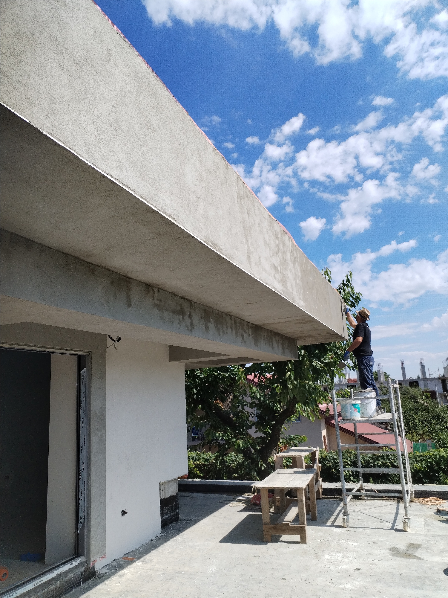 In Dumbrăveni, Romania, a person on a rolling scaffold is applying material to the underside of a concrete overhang. The individual is wearing a dark t-shirt, blue jeans, a straw hat, and blue gloves. They are holding a tool, possibly a trowel or brush, and appear to be working on a section of the concrete structure. Below the scaffold, two rustic wooden tables are placed on a concrete surface. The scene is outdoors, under a bright blue sky with scattered white clouds. In the background, green trees and buildings are visible, suggesting a residential or construction area. The overall impression is that of ongoing exterior building maintenance or construction work.