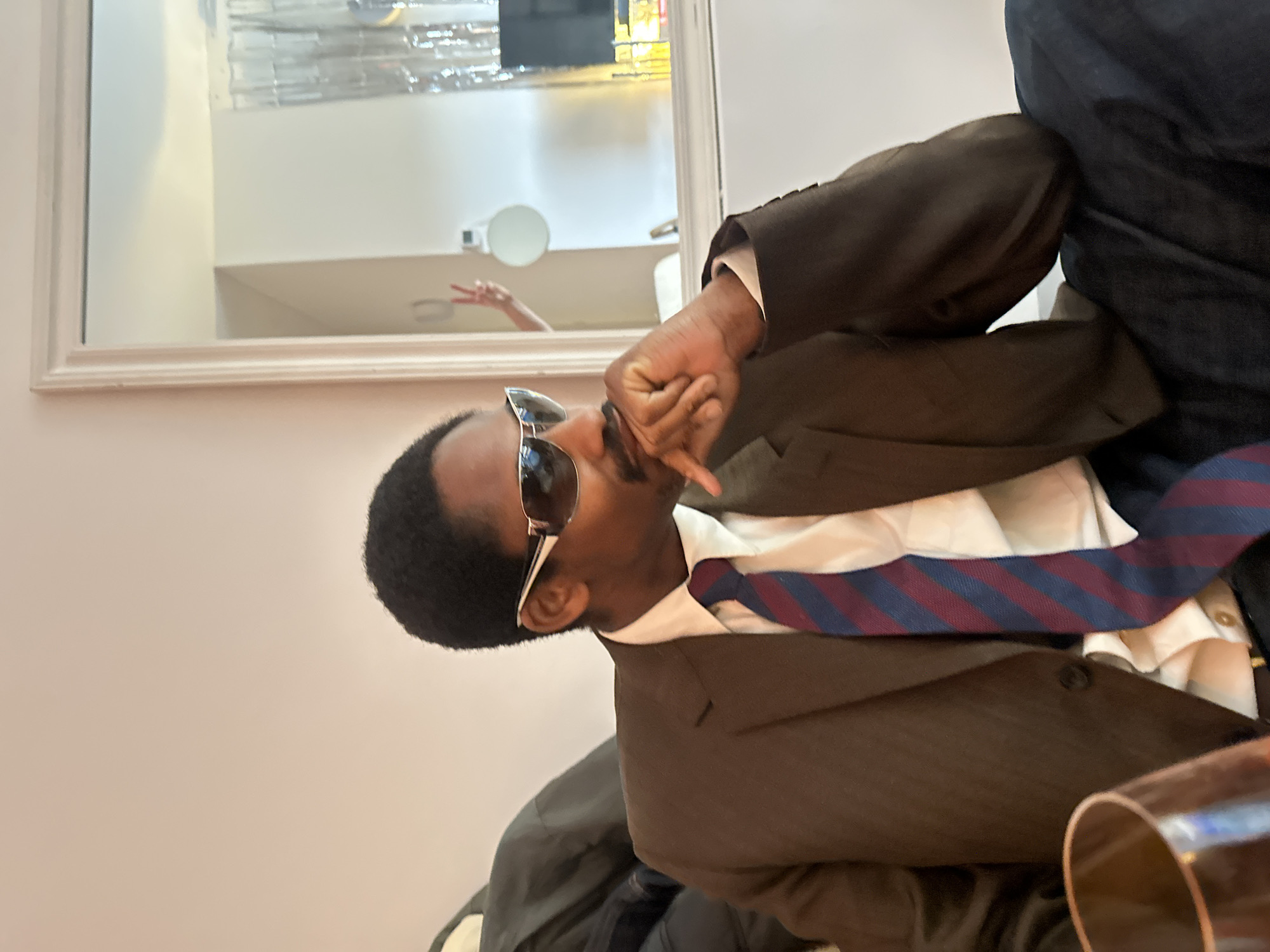 The image shows a man wearing sunglasses, a brown suit, and a blue and red striped tie. He is making a hand gesture, possibly a peace sign. A mirror in the background reflects a hand also making a peace sign. The location cannot be determined from the image.