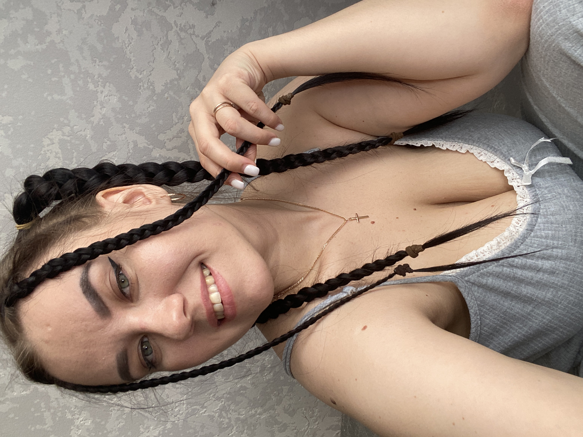 A fair-skinned woman with dark braided hair is shown in a close-up shot. She is wearing a grey, lace-trimmed top. She is smiling. She is also wearing a gold necklace with a cross pendant and a gold ring on her left ring finger. The background is a grey textured wall. The braids are tied with small brown hair ties at the ends. This scene is located in Chaikovsky, Russia.