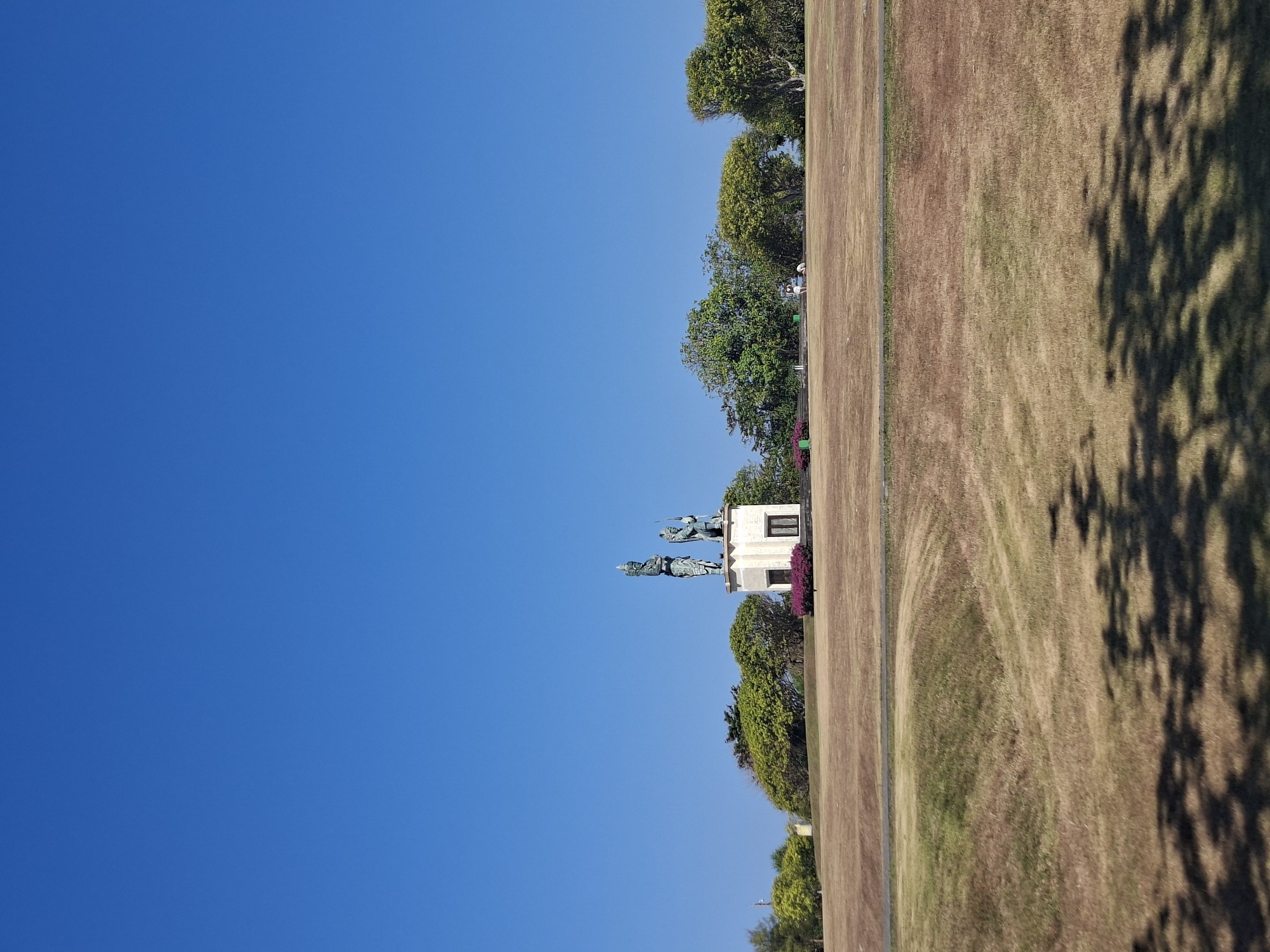 This image captures a daytime outdoor scene with a clear, bright blue sky dominating the upper portion. In the mid-ground, a statue of two figures stands atop a rectangular pedestal. The statue is the focal point of the scene, with the figures appearing to be made of a dark material, possibly bronze, and adorned with intricate details. The pedestal is lighter in color and features two windows and a doorway. In front of the pedestal, a strip of vibrant purple flowers adds a splash of color to the otherwise dry, golden-brown landscape.
The ground in the foreground and mid-ground is covered in dry, sparse grass, suggesting a warm or arid climate. Shadows of trees are cast across the foreground, indicating the presence of foliage out of frame. In the background, a line of lush green trees forms a natural barrier behind the statue. Further in the distance, on the horizon, a few structures are visible, and a small white object, possibly a car or a cart, is parked on the right side of the scene. There are also two people visible in the distance, on the right side, appearing to walk or stand in the grassy area.
The location is identified as Kuta Selatan, Indonesia. The overall impression is that of a public monument or memorial set within a park-like area, under a vast, cloudless sky.