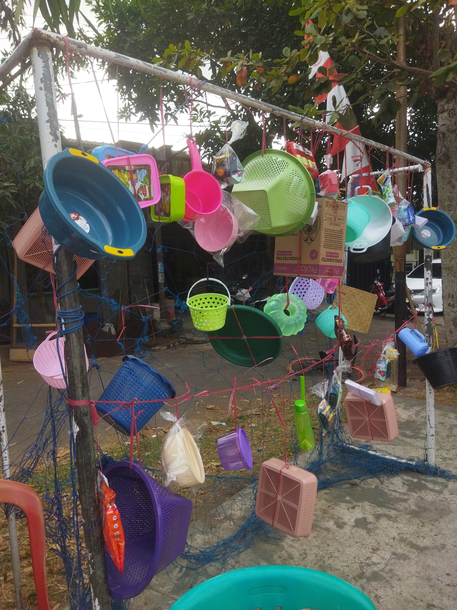 This image depicts an outdoor market stall located in the city of Banjar, Indonesia. The stall is set up along a roadside, with a variety of plastic household items, such as basins, colanders, buckets, and baskets, displayed on a metal frame. Many of these items are hung with red strings or attached to a blue net. Some of the plastic items are in bright colors, including blue, green, pink, and purple. There are also some packaged goods and bottles visible. In the background, there are trees, a white car, and what appears to be a building. The ground is paved with concrete and has some scattered leaves. The overall impression is that of a humble street vendor selling affordable plastic goods.
