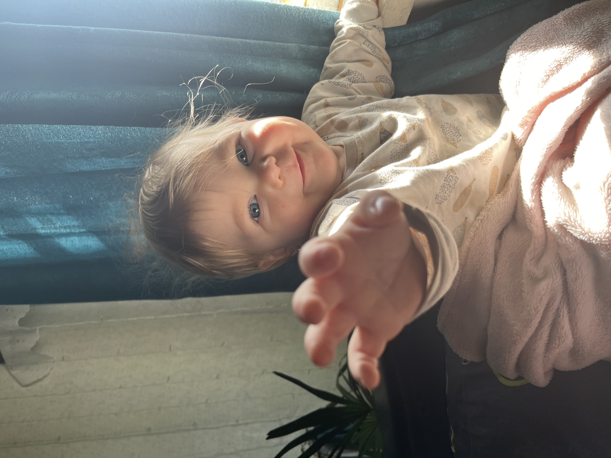 An overhead shot captures a smiling baby or young child with light blonde hair and bright blue eyes, lying on their back. The child, situated in an indoor setting in Фоки, Russia, wears a light-colored, long-sleeved onesie or pajamas adorned with a pattern of small, abstract shapes, possibly hedgehogs or feathers, in beige and grey tones. The baby's head is tilted, looking directly at the camera with a joyful expression, and one hand is playfully reaching forward, creating a sense of interaction.
The child rests on a dark teal or green textured fabric, likely a sofa or thick blanket. To the right, a soft, light pink fluffy blanket or textile is visible, heavily illuminated by strong natural sunlight coming from the upper right of the frame. This bright light also creates highlights on the child's hair and cheek, suggesting it is daytime. In the background, a light-colored wall with faint horizontal lines is visible, adding to the domestic feel of the scene. A glimpse of a green potted plant can be seen in the lower part of the frame. No text is visible in the image.