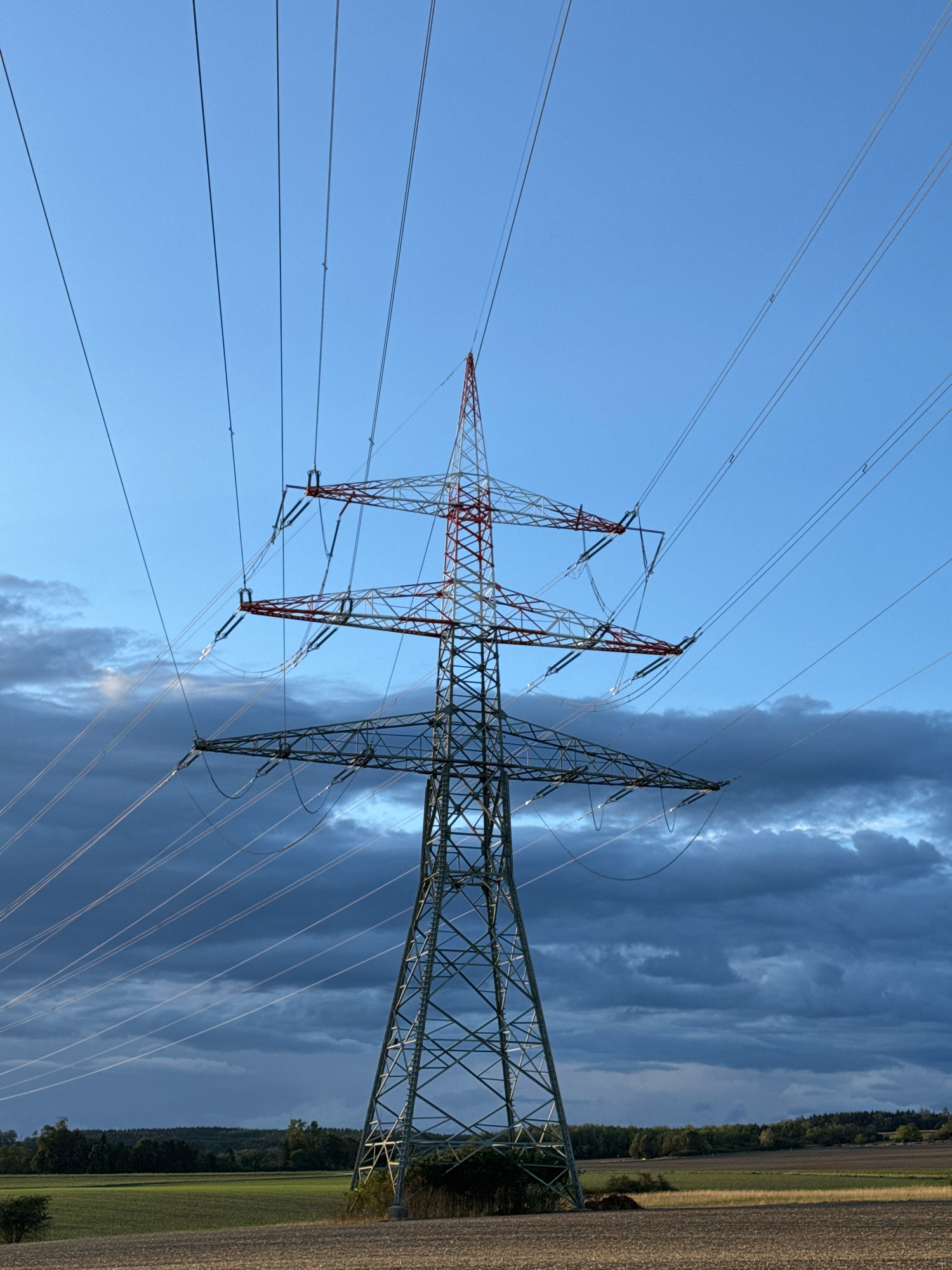 A tall, lattice-style electrical tower stands prominently in the foreground against a gradient sky of clear blue near the top and darker, cloud-filled hues towards the horizon. The tower is constructed of metal and has multiple crossarms from which numerous power lines extend outwards, disappearing into the sky. The upper section of the tower is painted in red and white bands, possibly for visibility. Below the tower, a field with dry, brown soil is visible, transitioning into green agricultural land dotted with trees in the middle ground and a dense treeline in the background. The overall scene suggests an outdoor rural environment with a clear sky and some cloud cover. The image was taken in the municipality of Wettstetten, Germany.