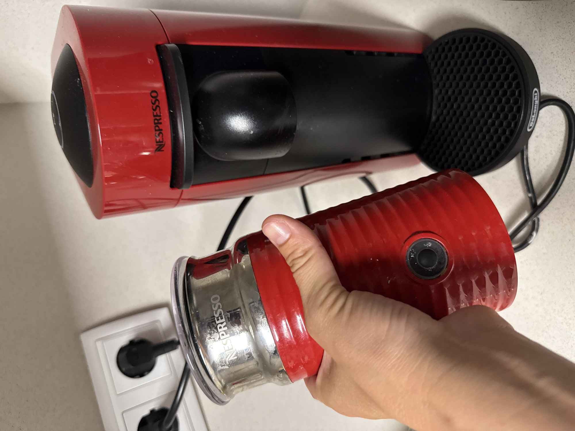 In Puig-reig, Spain, a Nespresso coffee machine and a Nespresso milk frother are displayed on a countertop. The red Nespresso coffee machine, positioned on the right, features a black spout and a removable drip tray with a honeycomb pattern. A white electrical outlet with two black plugs is visible on the left side of the image. A hand is holding the red Nespresso milk frother, which has a metallic lid inscribed with the Nespresso logo. The scene appears to be an indoor domestic setting.