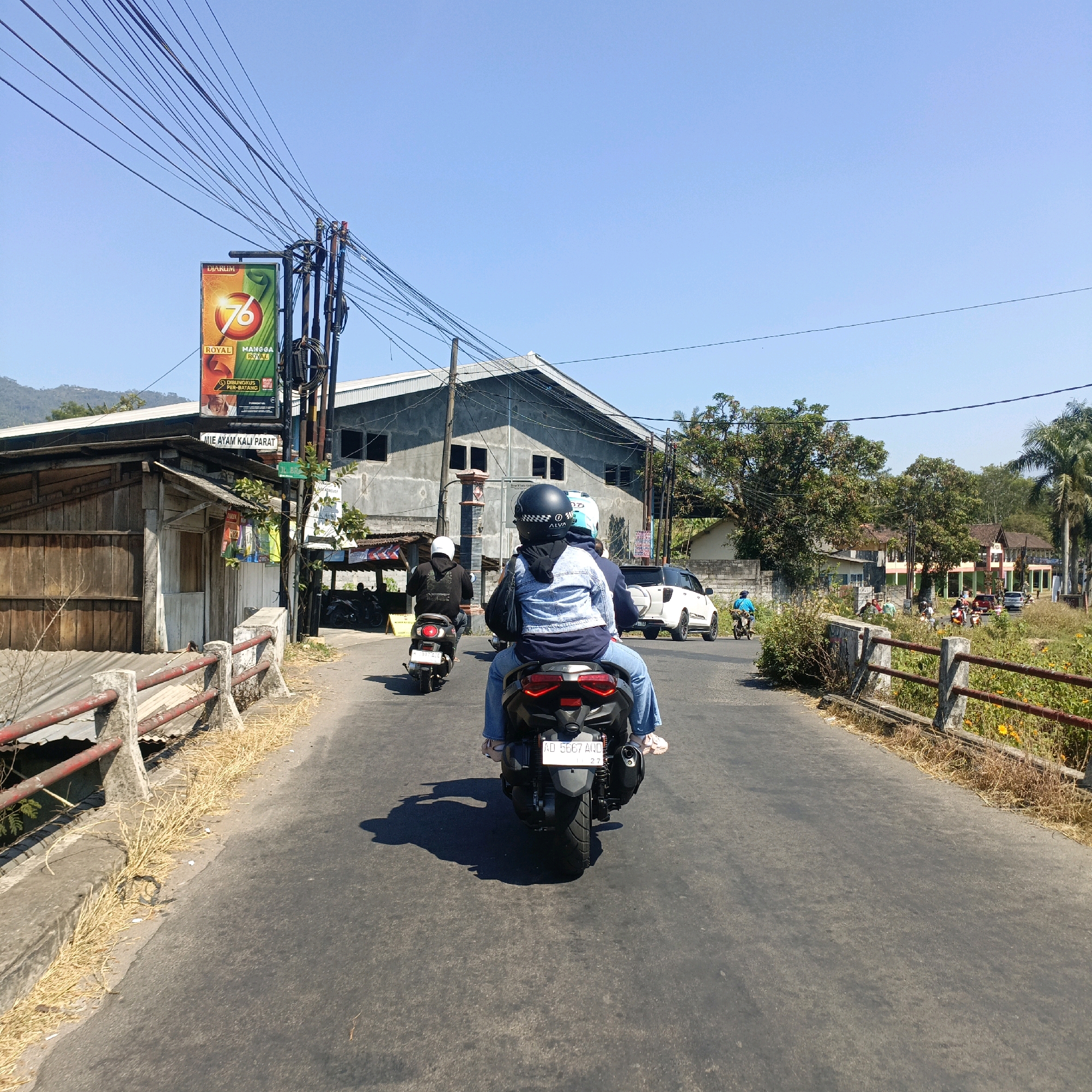 The image depicts a street scene in Kaliwangi, Indonesia, under a clear blue sky. A motorcycle with two occupants, viewed from behind, is the central subject, traveling down a paved road. The driver wears a helmet and a denim jacket, and the passenger in the back is also wearing a helmet and a blue jacket with light blue jeans. The motorcycle has a license plate visible. Other vehicles, including a white SUV and another motorcycle with a single rider, are present further down the road. On the left side of the road, there is a weathered wooden building with a corrugated metal roof, and a billboard advertising "DIARULM 76 ROYAL MANGGA ROYAL." A street sign that reads "MIE AYAM KALI PARAT" is also visible. A concrete and red-railed bridge or embankment lines the left side of the road, with dry grass and debris along its edge. The right side of the road features a more developed area with buildings, trees, and more vegetation. The overall impression is a sunny, daytime scene in a semi-urban or rural Indonesian setting.