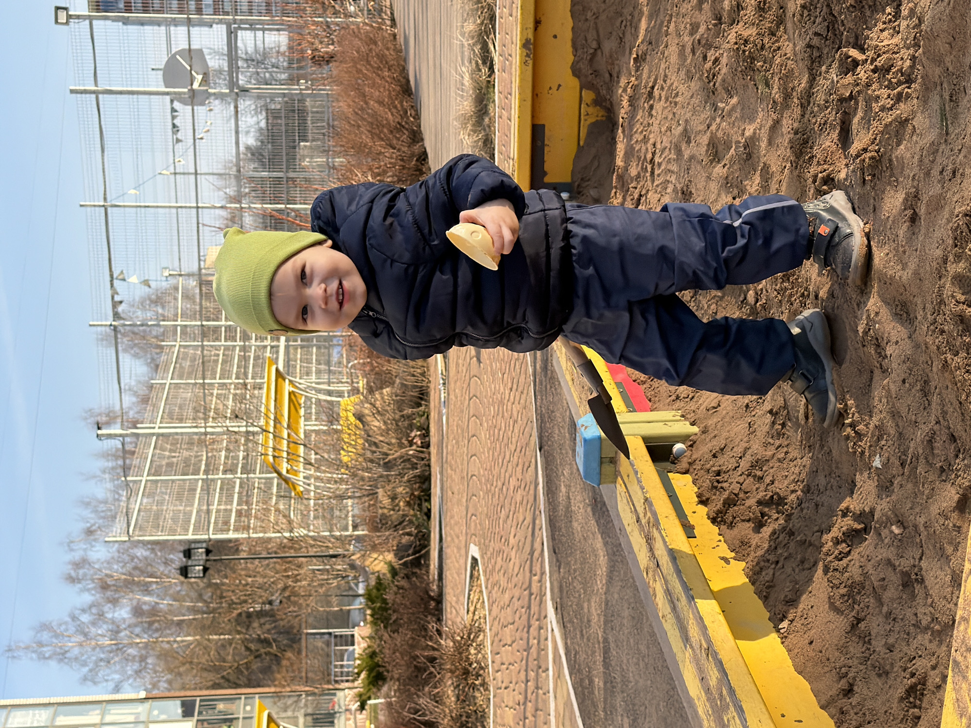 A young child, appearing to be approximately 2 years old, stands in a sandbox. The child wears a dark blue padded jacket, matching dark blue trousers with light gray stripes near the cuffs, gray boots with black straps, and a light green knitted hat. The child holds a small, light yellow object in their hand.
The sandbox is constructed with yellow wooden planks and contains sand. The ground around the sandbox consists of sand and dirt. Behind the sandbox, a brick walkway with a decorative patterned surface leads to a building.
In the background, there is a metal fence structure, trees, and a building with light-colored walls and windows. The sky is light blue.
Based on visual clues and potential local knowledge, the location is likely a playground or recreational area in the city of Izhevsk, Russia.