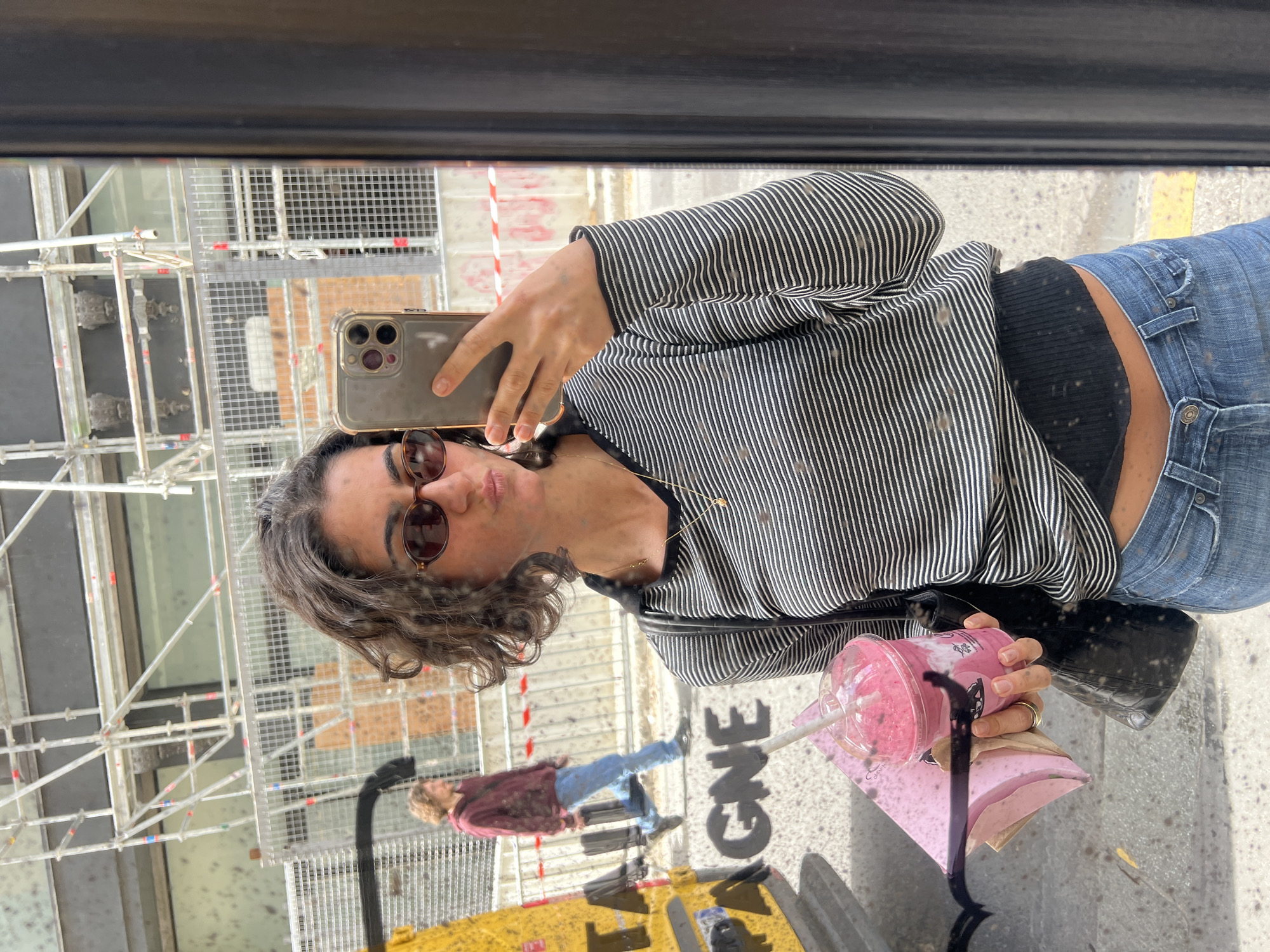 This is a mirror selfie showing a woman outdoors in an urban setting. The image is taken through a reflective surface, likely a shop window or large mirror, and is oriented vertically in the provided file, so the subjects appear on their sides.
The main subject, a woman with dark, curly hair, wears dark sunglasses, a black and white striped long-sleeved top, a black bralette, and blue jeans. She is holding a smartphone with a clear case in her left hand to take the selfie, and a clear plastic cup with a pink beverage and a straw in her right hand. A gold ring is visible on her finger, and a thin gold necklace hangs around her neck. She has a slight pursed-lip expression. A black shoulder bag is visible on her right side.
The background, reflected in the mirror, includes extensive metal scaffolding, suggesting a construction site or building renovation. Another person, a man wearing a reddish-brown top and blue jeans, is visible in the distance, reflected in the mirror. The ground features a yellow curb or barrier, and the word "ONE" is visible in large, dark letters on this surface. Various specks and dust particles are noticeable on the reflective surface, covering parts of the woman's clothing and the ground. The scene appears to be taking place during the daytime under bright conditions. The location context indicates this is likely a street in Paris, France.