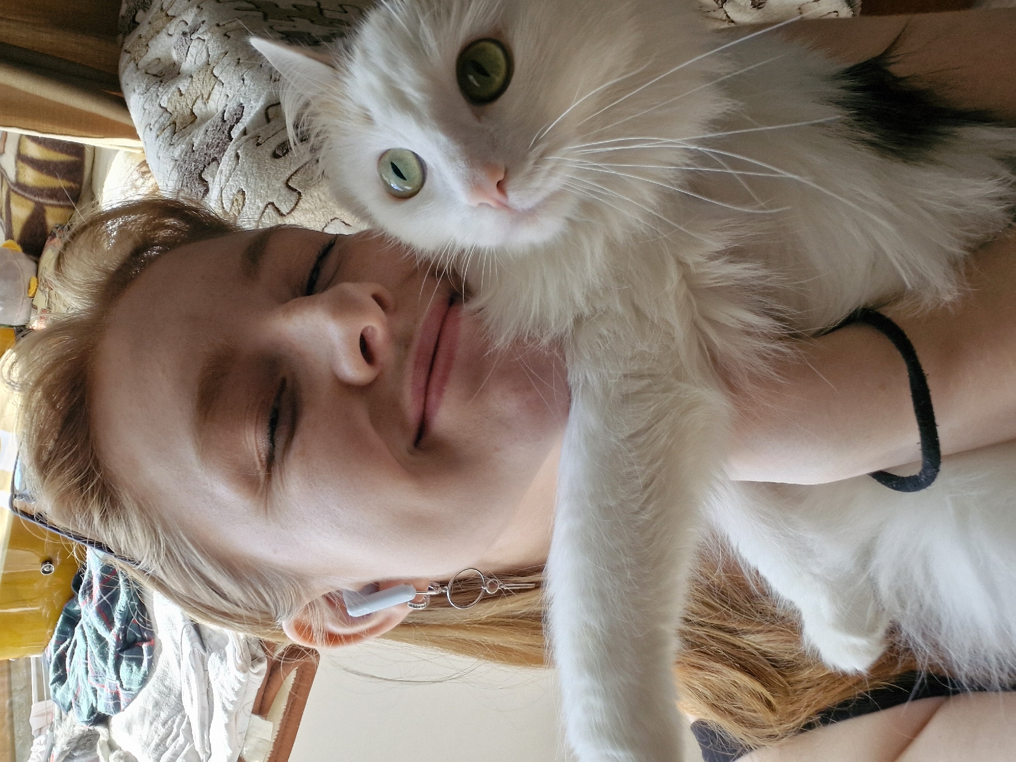 A woman with fair skin and reddish-blonde hair lies on her side, her eyes closed. She is wearing an earring on her right ear and what appears to be a wireless earbud in her left. She is holding a white cat with green eyes. The cat has some black fur on the top of its head. The background is cluttered with objects including what appears to be clothing, a bedspread, and other miscellaneous items. The location cannot be determined to be Yekaterinburg, Russia without more information.
