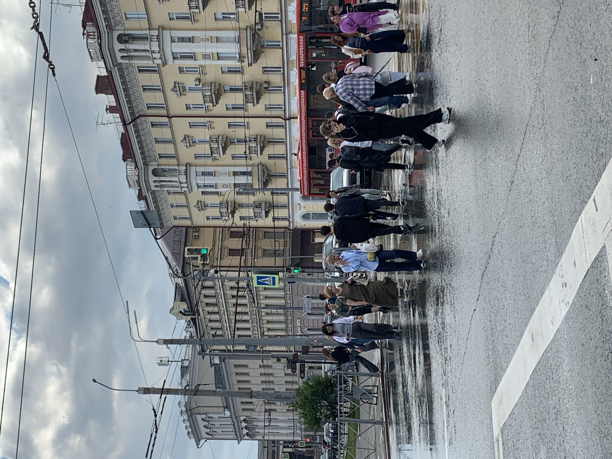 This media file depicts a street scene in Kazan, Russia, likely during the daytime, under overcast skies and after a period of rain, as evidenced by the wet pavement and reflections. The image is oriented vertically, with the bottom of the frame showing a crosswalk.
The primary subjects are a group of pedestrians crossing a street. They are diverse in age and attire, with some dressed in casual clothing like jeans, t-shirts, and jackets, while others wear more formal or stylish outfits. Their expressions are not clearly discernible due to the distance and resolution, but the overall atmosphere appears to be one of everyday urban activity.
The background features a street lined with multi-story buildings with ornate architectural details, characteristic of European cities. These buildings appear to be a mix of residential and commercial establishments. Visible elements include windows, balconies, and some air conditioning units. There are also streetlights and traffic signals, including a green traffic light visible in the upper portion of the image, suggesting that traffic is permitted to move. Power lines and tram wires are also prominent, indicating an active public transportation system.
A red tram with Cyrillic lettering can be seen in the middle of the frame. Though the text is difficult to read clearly, the presence of Cyrillic script confirms the Russian location. A pedestrian crossing sign with a pictogram of a person walking is also visible. The wet street surface reflects the surrounding buildings and the sky, adding a dynamic quality to the scene. The overall impression is of a bustling urban environment engaged in its daily routine, slightly subdued by the weather.