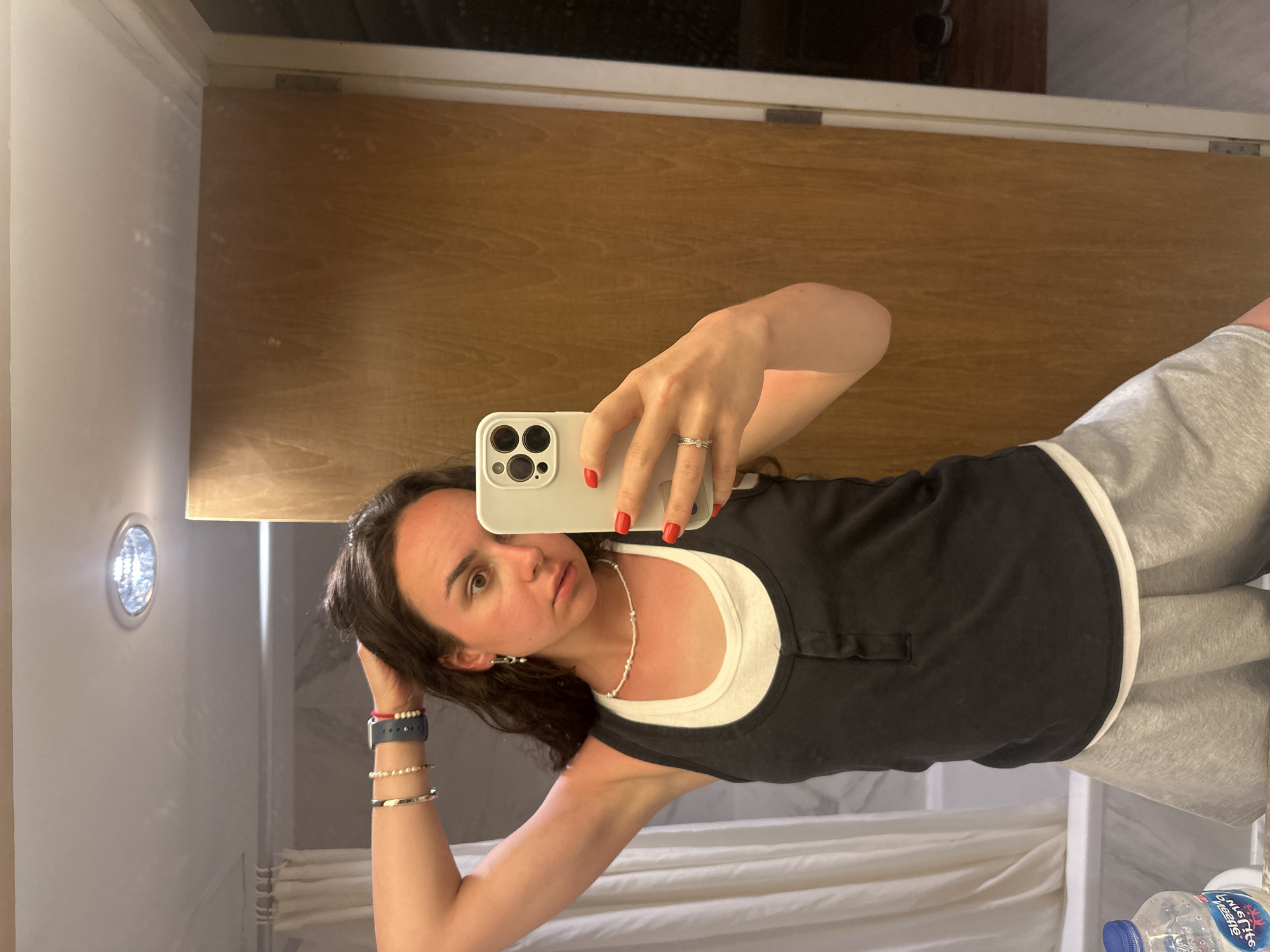 A young woman is taking a selfie in a bathroom, facing a mirror. She holds an iPhone 13 or later generation phone in her right hand, her fingers with red nail polish. The woman is wearing a black tank top over a white tank top, and grey sweatpants. She has dark hair, earrings, a white bead necklace, and bracelets on her left wrist. The bathroom is illuminated by a recessed ceiling light. A closed wooden door is visible behind her, as well as a shower or bathtub with white curtains. There is a water bottle visible at the bottom right corner. The location is Pattaya City, Thailand.