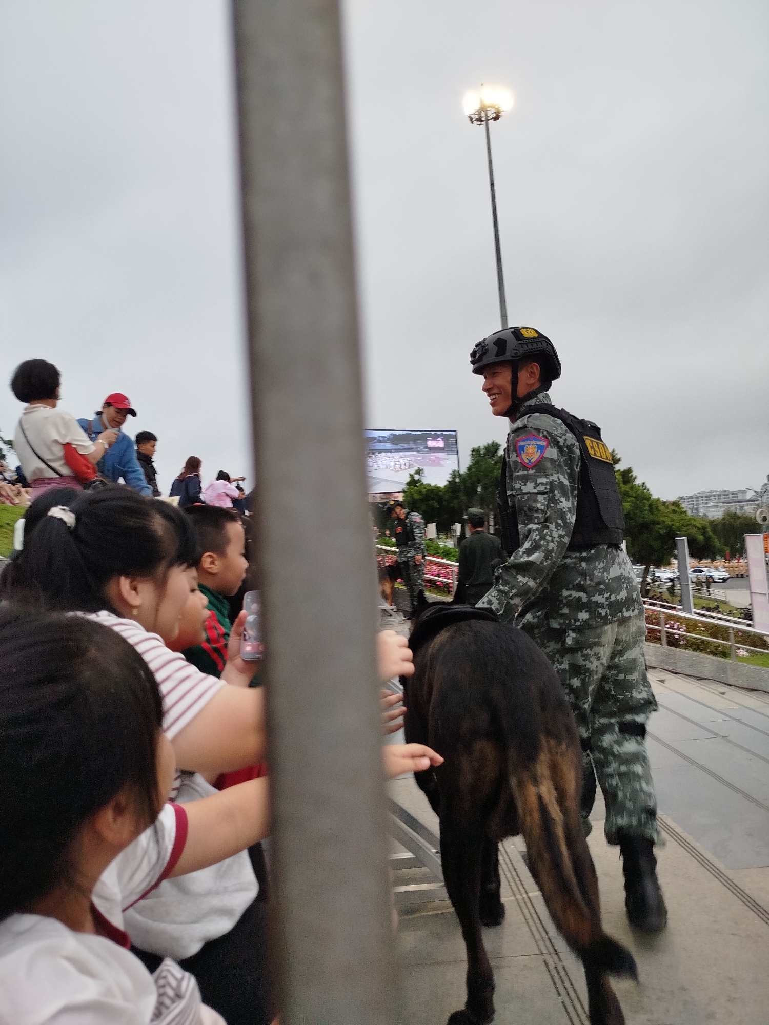 A male police officer in a camouflage uniform and tactical helmet is walking down a paved pathway with a German Shepherd police dog. The officer is smiling and looking to his left. He has a patch on his left shoulder and the letters "CSM" on his vest. Behind him, several people, including children, are standing and watching. The scene appears to be outdoors with a cloudy sky. In the background, there are trees, buildings, and streetlights. The pathway is lined with railings and flower beds. This scene could be in the city of Da Lat, Vietnam, as the architecture and vegetation are consistent with the region, though this cannot be definitively confirmed from the image alone.