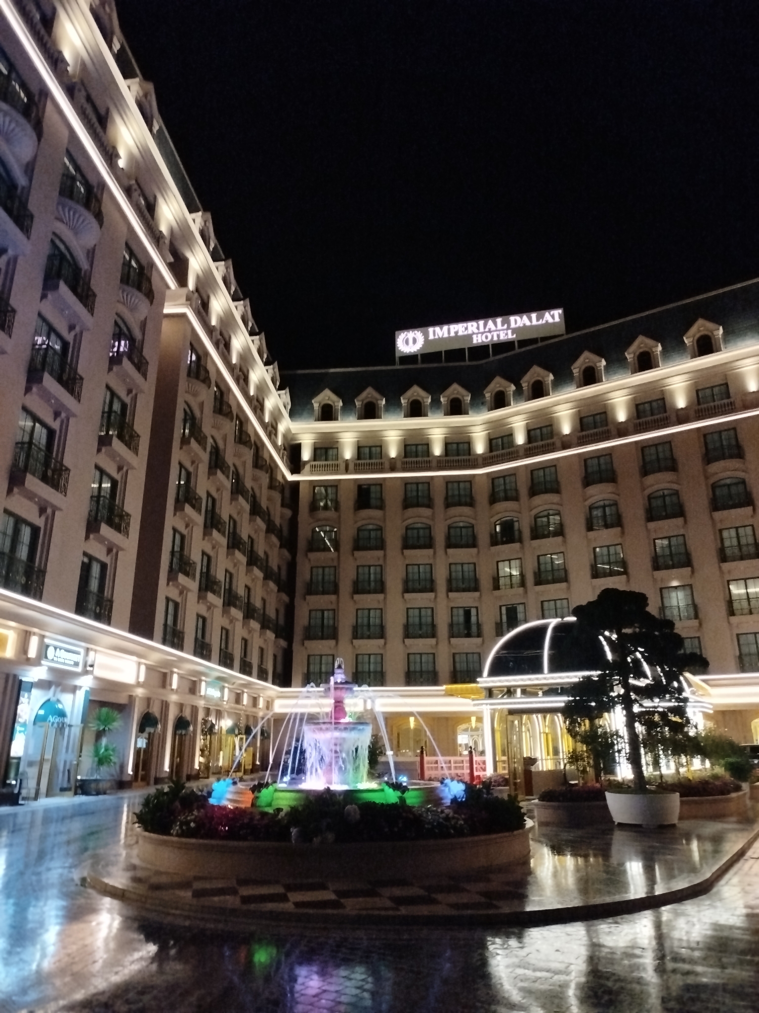 At night, the Imperial Dalat Hotel in Lâm Viên - Đà Lạt, Vietnam is captured with its fountain illuminated in various colors. The hotel's architecture features beige walls with dark windows and roofline decorations. A sign reading "IMPERIAL DALAT HOTEL" is visible on the roof. The fountain, situated in a circular garden bed, displays a range of colors including blue, green, and pink. The ground is reflective, indicating a wet surface. The scene is devoid of human presence.