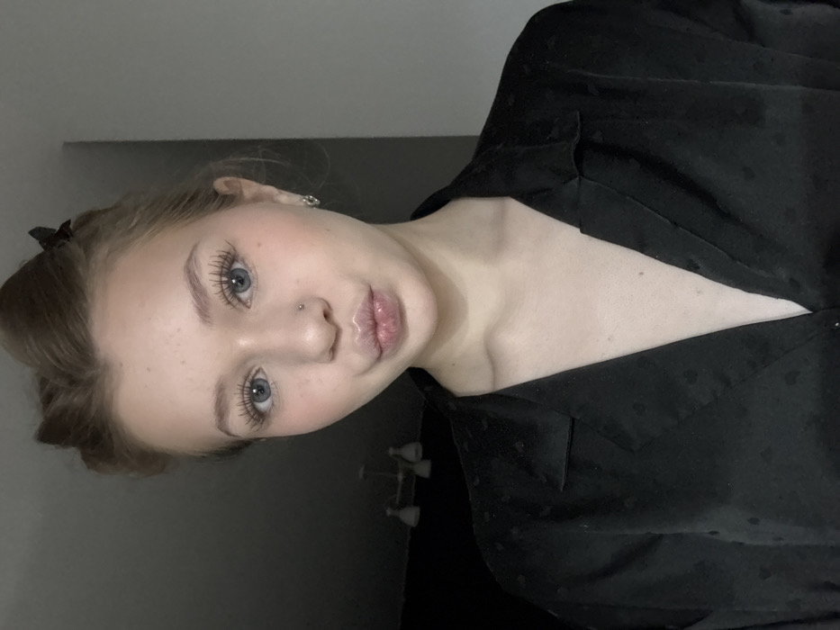 The image depicts a young Caucasian female in a posed self-portrait, captured indoors in Yekaterinburg, Russia. Her head and upper torso are visible. Light-colored hair, possibly blonde, is styled with the front sections swept back and held by a small, dark butterfly-style claw clip on the viewer's left. Her blue eyes feature notably long eyelashes. A small, silver-toned stud is visible in her left nostril, and small silver hoop earrings adorn her ears. Her lips are pursed and appear glossy, forming a "kissing" expression. She wears a black, collared shirt or blouse with a subtle, repeating pattern of small, dark hearts. The garment is unbuttoned to reveal the upper sternum area. The background is out of focus and dimly lit, indicating an indoor setting. A multi-bulb ceiling fixture is partially visible on the left against a dark wall, while a lighter, neutral-toned wall occupies the right side of the frame.