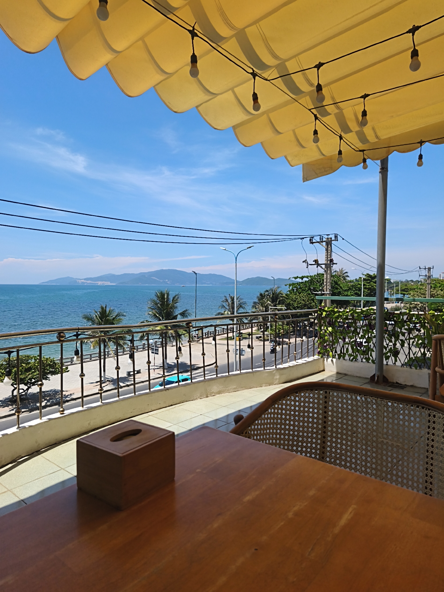 The image captures a view from a balcony or patio overlooking a coastal scene in Bac Nha Trang Ward, Vietnam. In the foreground, a wooden table is visible, with a wooden tissue box placed on it. To the right, the back of a wicker chair is partially visible. The balcony railing, made of metal with circular decorative elements, separates the foreground from the middle ground. Beyond the railing, a palm-lined promenade runs alongside a road with some traffic. The sea is a prominent feature, with calm blue water stretching towards a distant mountain range under a clear blue sky with scattered clouds. The scene is partially shaded by a yellow, scalloped awning overhead, with decorative string lights attached to it. Electrical poles and wires are visible in the background. The overall impression is a relaxed, seaside ambiance.