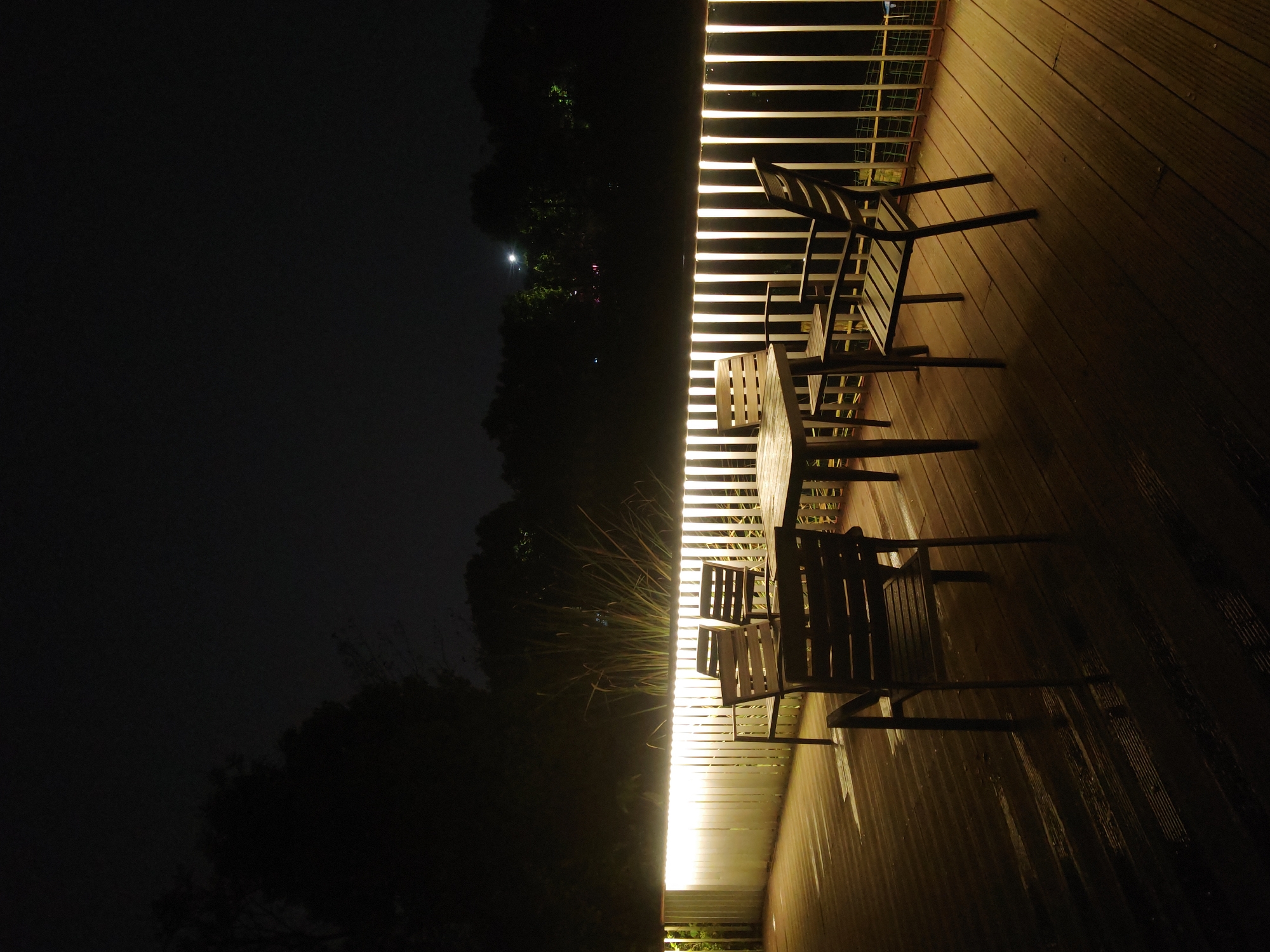 The image depicts an outdoor dining area in Chongqing High-tech Zone, China, at night. A wooden deck is illuminated by a linear light source along a railing. The railing has vertical bars. A wooden table and four chairs sit on the deck. Beyond the railing, there are dark trees and a hint of faint light in the distance. The left side of the image is dominated by darkness.