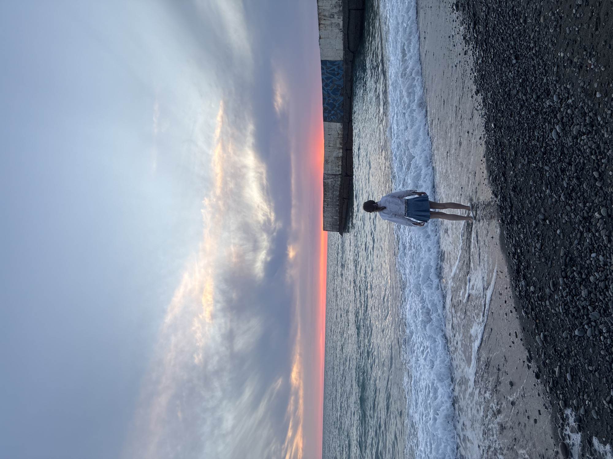 A person stands facing a body of water, with waves washing onto a pebble beach. The person is wearing a white shirt and a blue skirt, and their feet are submerged in the shallow water. In the distance, a concrete breakwater extends into the sea. The sky is filled with clouds, and the sun appears to be setting, casting a warm orange glow on the horizon. The scene is likely captured at a beach in Sochi, Russia, given the prominent breakwater which is characteristic of the city's coastline.