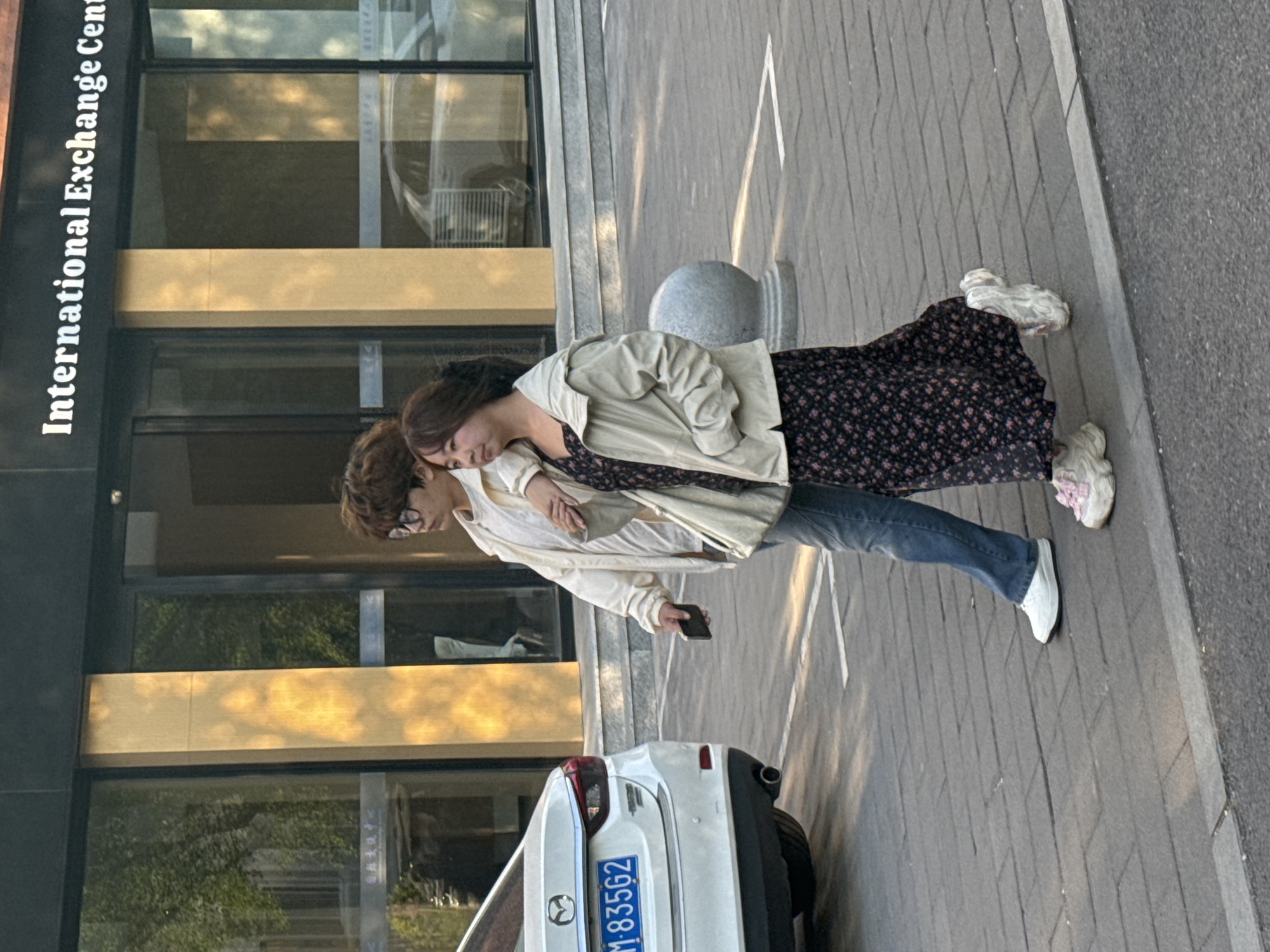 The photograph captures two individuals standing on a gray brick-paved area adjacent to a building labeled "International Exchange Center." The building's facade features windows reflecting sunlight and nearby greenery. A white vehicle, partially visible, bears a license plate that reads "M·835G2."
One person, wearing glasses, is positioned holding a mobile phone. The other leans on them. The environment appears to be a city street, possibly in Zhongtun, China, as suggested by the building signage.