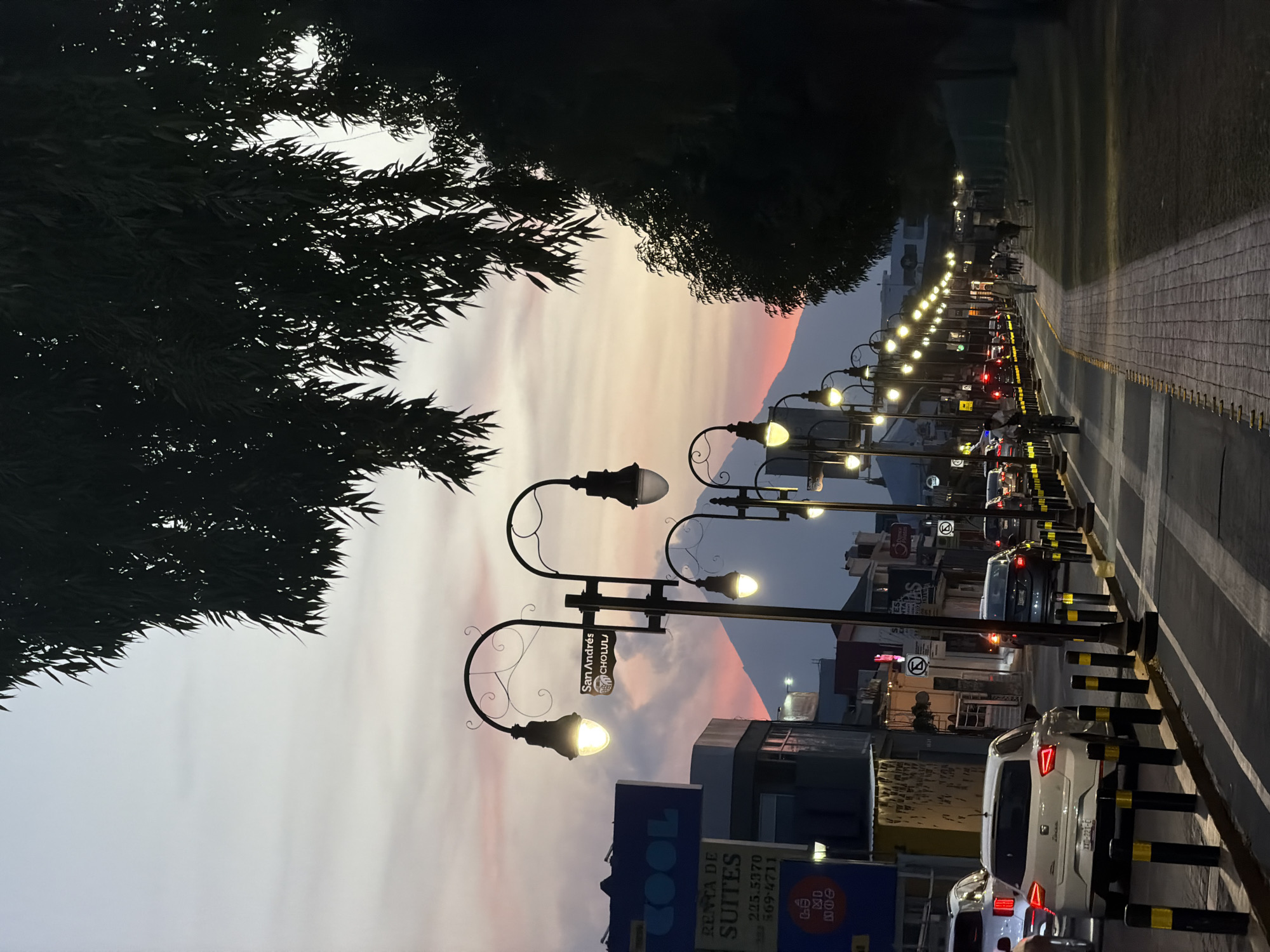 The image captures a vibrant twilight scene on a street in San Andrés Cholula, Mexico. The sky, a dominant feature, displays soft pastel hues of light blue, grey, pink, and orange, indicating either dawn or dusk. In the distance, a majestic mountain or volcano, likely Popocatépetl or Iztaccihuatl, is silhouetted against the colorful sky, with its peak catching the last rays of sunlight in a delicate pink glow.
Along the left side of the frame, dark, dense foliage of trees is silhouetted against the sky, adding depth to the composition. The right side of the image features a paved street lined with ornate streetlights that emit a warm, yellow light, illuminating the area as daylight fades. Several cars are visible on the road; some appear to be parked, while others suggest evening traffic, with their headlights and taillights glowing. A distinctive sidewalk or pedestrian path, paved with bricks and bordered by black and yellow bollards, runs parallel to the street on the far right.
Low-rise buildings line the street, featuring various commercial establishments. A large blue sign prominently displays the word "COOL" and advertises "RENTA DE SUITES" with phone numbers "222.5370" and "569-4711." Crucially, a street sign attached to one of the decorative lampposts clearly reads "San Andrés CHOLULA," confirming the location. The scene conveys a sense of a busy urban environment transitioning into evening, with the natural beauty of the sunset and distant mountain providing a stunning backdrop.