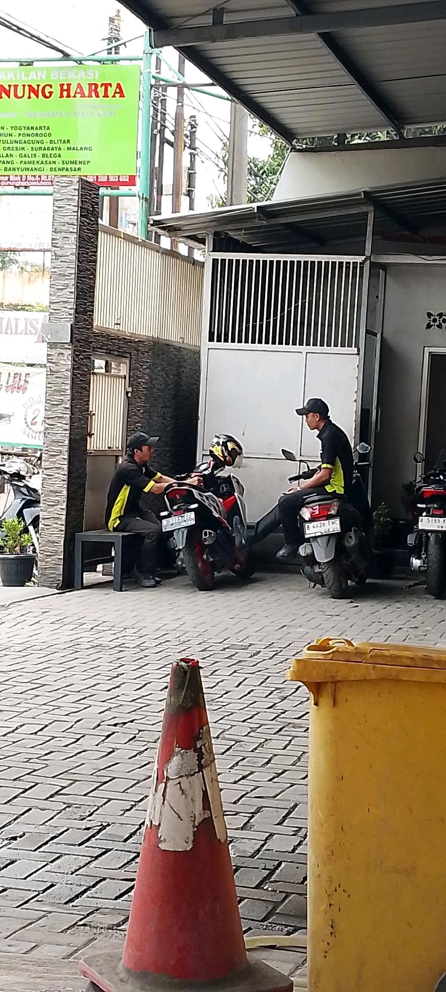 Two men in yellow and black uniforms with black caps are sitting on motorcycles in front of a building in what appears to be a commercial or service area. The man on the left is sitting on a red and black motorcycle, leaning forward with his hands on the handlebars. The man on the right is sitting on a gray motorcycle, looking to his left. Both men appear to be waiting. In the foreground, a red and white traffic cone stands on a paved walkway. Behind the men and motorcycles, a building with a metal roof and a white gate is visible. A green sign with text in Indonesian, indicating "AKILAN BEKASI GUNUNG HARTA SOLUSI," is mounted on a pole to the left of the building. This sign suggests the location is in Bekasi, Indonesia. The overall environment appears to be an outdoor, paved area in front of a business.