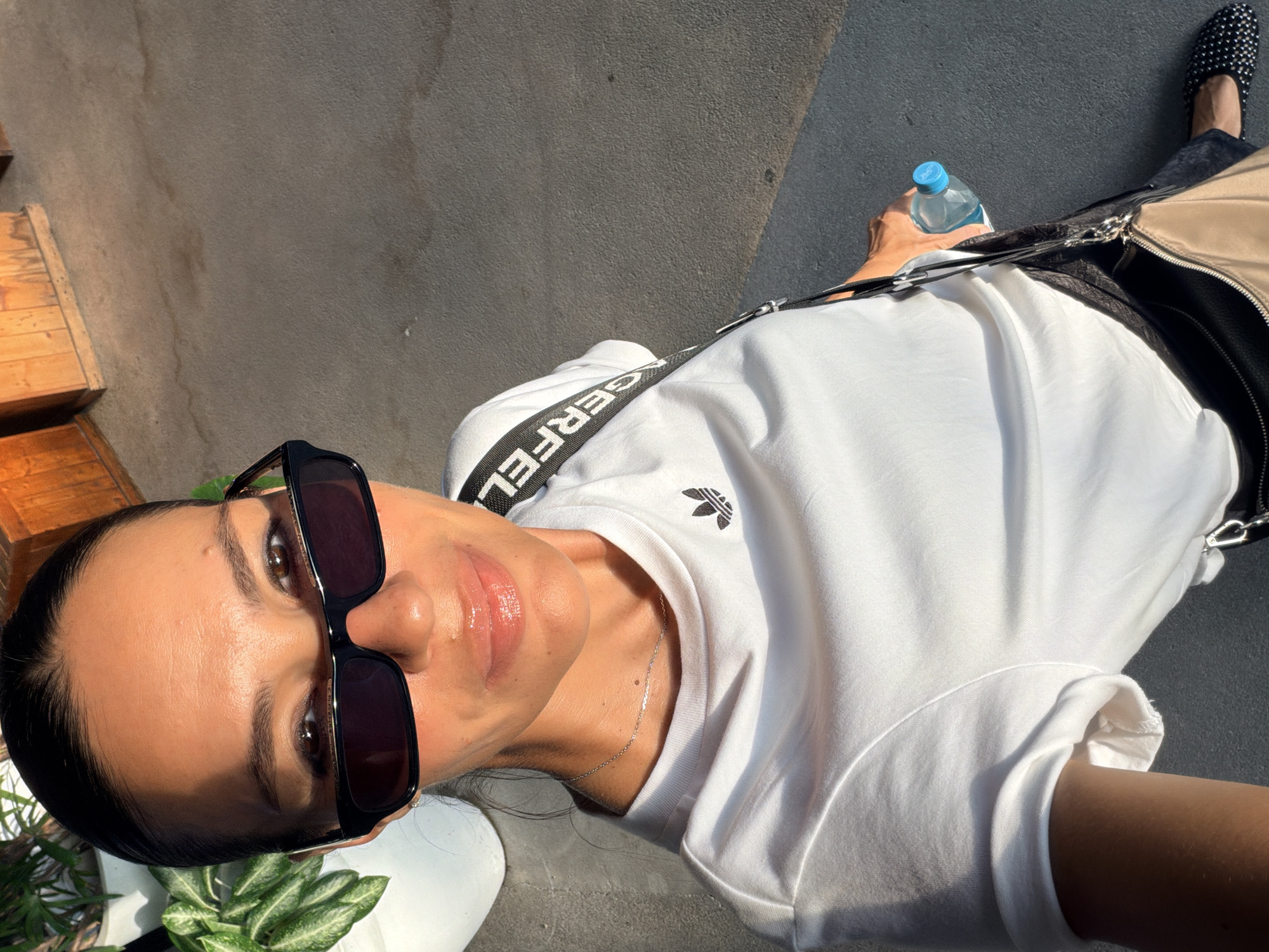 A woman is taking a selfie outdoors, wearing a white t-shirt with an Adidas logo, black sunglasses, and a silver necklace. She has on a black skirt, a light brown purse, and black studded flats. In her right hand, she is holding a bottle of water. The background shows a textured gray surface, possibly a sidewalk or pavement, with wooden structures visible in the distance. The lighting suggests it is daytime, with sunlight casting shadows and highlighting parts of the scene. The environment appears to be urban or a public outdoor space. There is no specific information in the image to indicate the location is in Ko Samui District, Thailand.