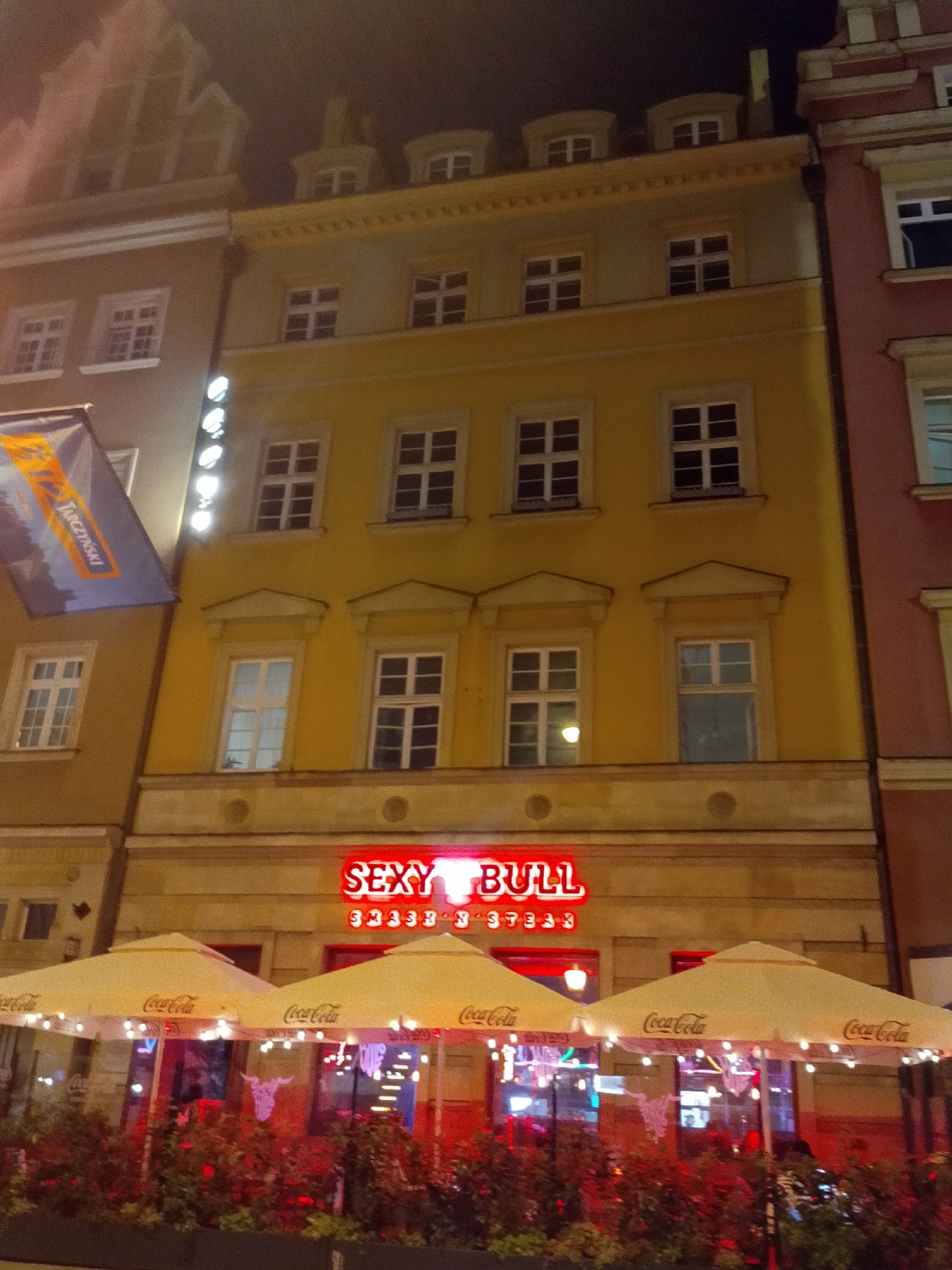 The image shows a nighttime view of a restaurant called "SEXY BULL" in Wrocław, Poland, situated on a street with multiple buildings. The restaurant features bright red neon signage and is partially obscured by two large, cream-colored umbrellas advertising "Coca-Cola." The umbrellas are set up over outdoor seating areas, which are decorated with red lights and potted plants in the foreground. Behind the umbrellas, the interior of the restaurant is visible through large windows, revealing more red lighting and some decorative elements, including what appears to be a steer's skull. The buildings surrounding the restaurant are multi-story structures with numerous windows. One building to the left has a blue banner with yellow text that reads "TARZYŃSKI." The lighting is dim, characteristic of a nighttime city scene, with the neon sign of the restaurant being a prominent light source.