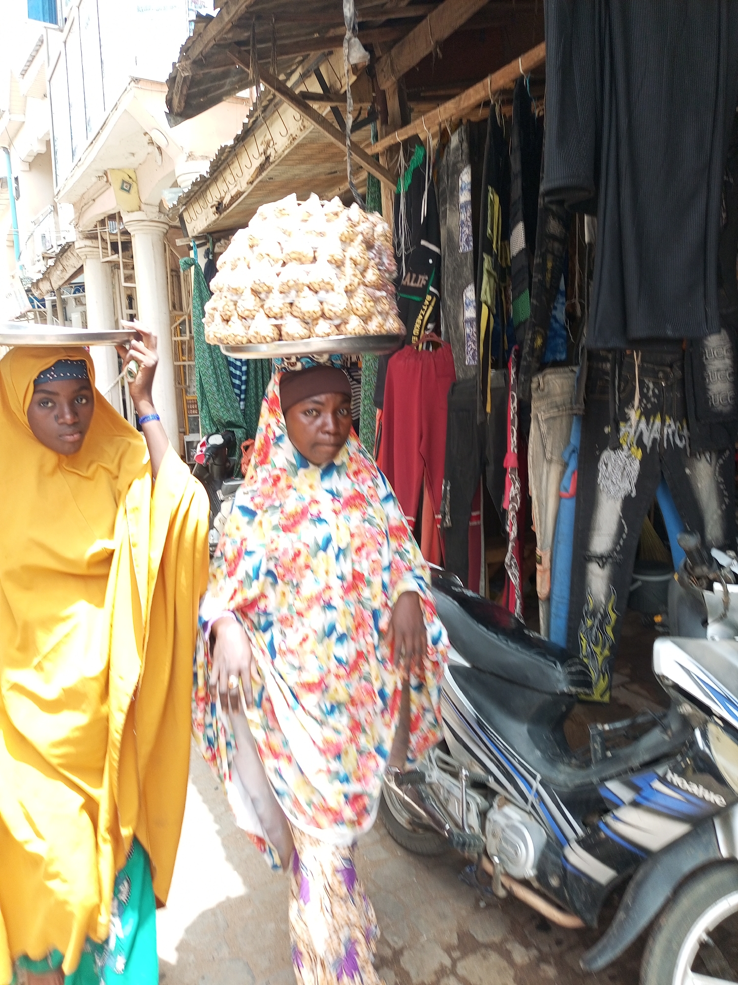 In Arawa, Nigeria, two women are walking on a street. The woman on the right is carrying a large stack of bread rolls balanced on a tray on her head. She is wearing a floral patterned headscarf and gown. The woman on the left has a tray over her head and is wearing a yellow headscarf and gown. Behind them, clothing hangs for sale on a makeshift wooden rack. A parked motorcycle is visible. The building architecture suggests a commercial district.