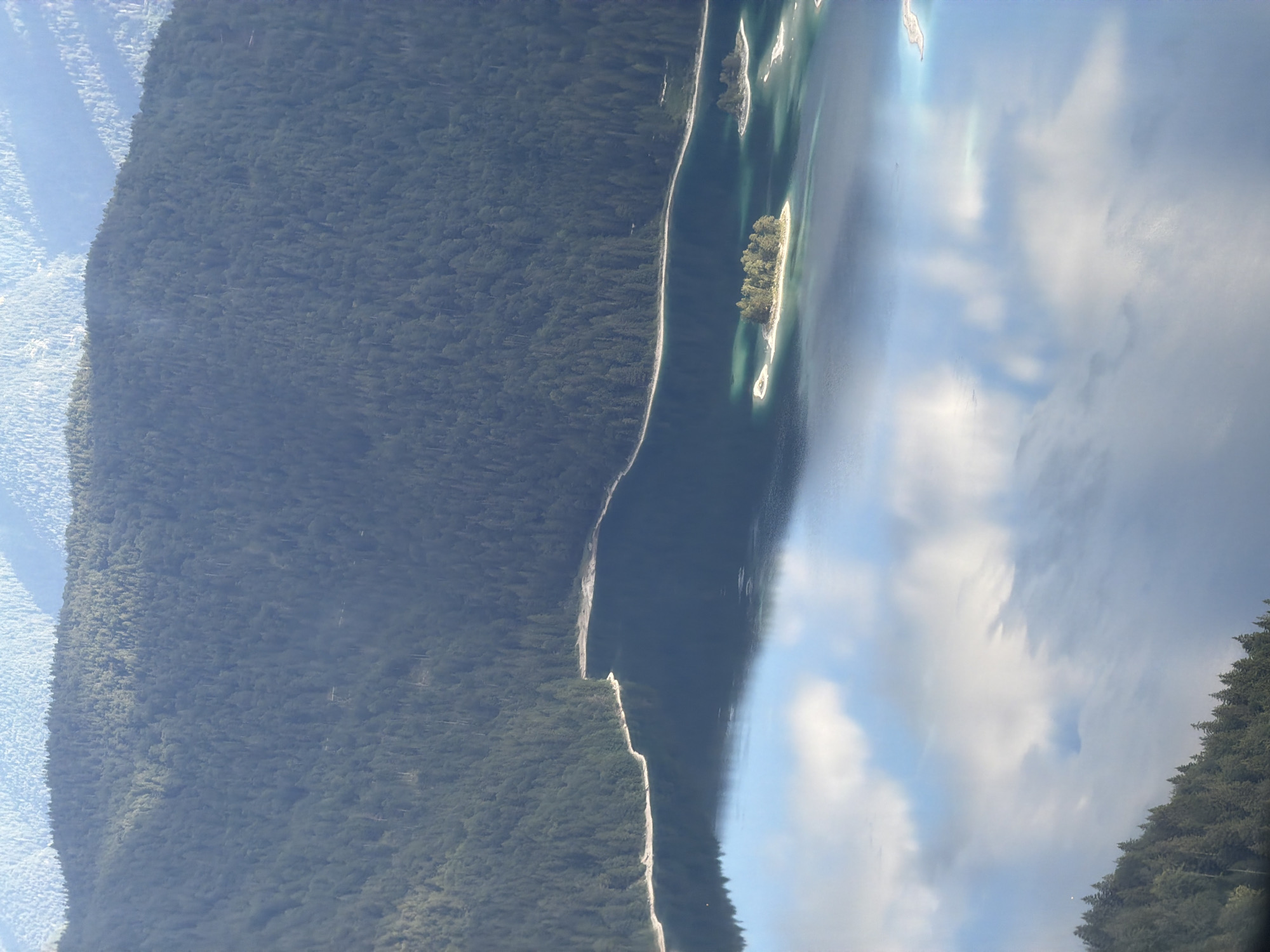 An aerial view captures a picturesque landscape in Grainau, Germany, dominated by a serene lake and verdant mountains. The Eibsee, known for its clear turquoise waters, is prominently featured. Several small, tree-covered islands dot the lake, adding to its charm. The surrounding mountains are densely forested, with a mix of dark green conifer trees creating a rich tapestry of color. The sky is partially overcast, with fluffy white clouds reflecting on the still surface of the lake, creating a mirror-like effect. The image conveys a sense of tranquility and natural beauty.