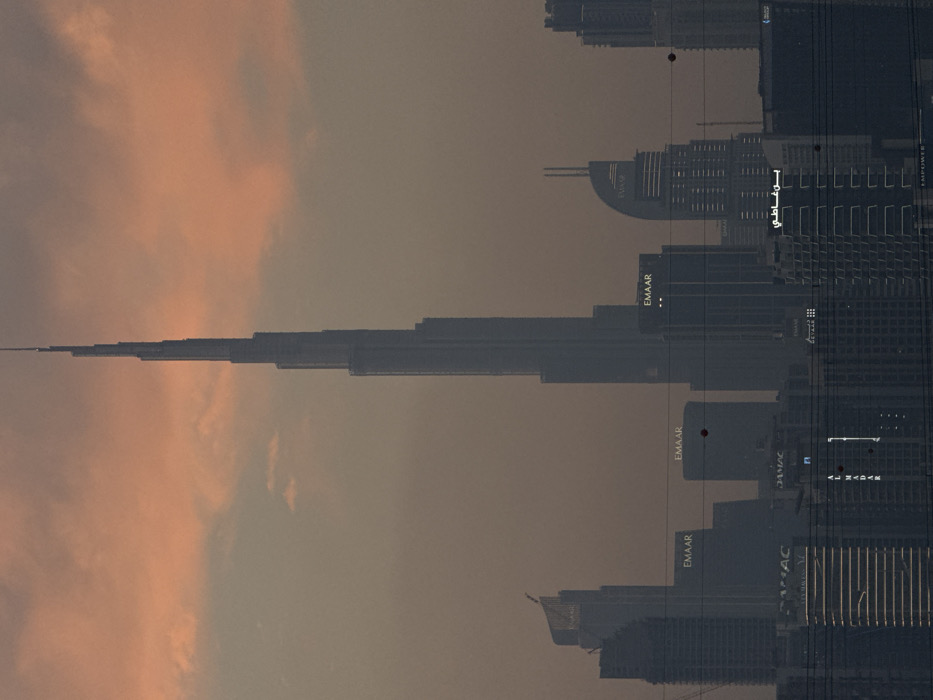 The image presents a hazy, long-distance view of the Dubai skyline in the United Arab Emirates, at either dawn or dusk. The dominant subject is the Burj Khalifa, a prominent skyscraper centrally positioned, extending towards the upper edge of the frame. Its tapered, multi-tiered structure is rendered in a dark, silhouetted tone against an overcast sky featuring diffuse orange and pink clouds.
Below the Burj Khalifa and extending across the midground and foreground are numerous other high-rise buildings of varying architectural styles and heights. Several buildings display illuminated "EMAAR" branding. Other visible building names include "AL MADAR," "DAMAC," "EMPOWER," and Arabic text such as "بن فاضلي".
Multiple dark, thin horizontal lines, consistent with utility wires or cables, traverse the lower two-thirds of the image, partially obscuring the buildings. Scattered across these wires and within the sky are small, dark, and a few reddish-orange circular or irregular specks. No individuals, vehicles, or specific actions are discernible within the frame. The overall scene is characterized by soft, diffused lighting and a pervasive atmospheric haze.
