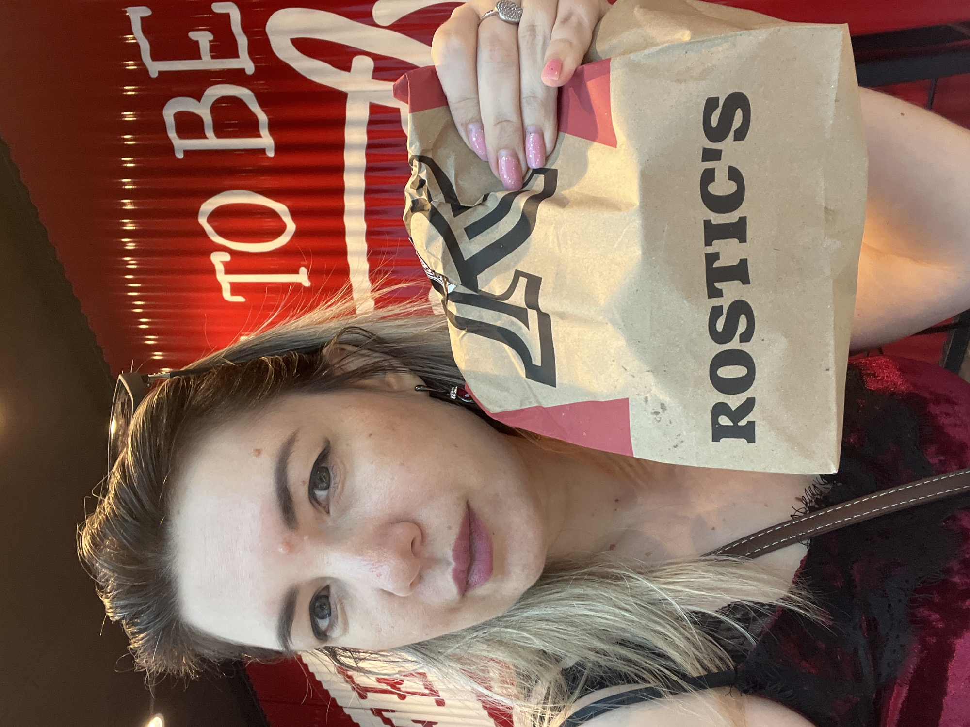 The image shows a woman holding a brown paper bag with "ROSTIC'S" written on it. She is wearing a dark red velvet top with lace trim. The background is a red corrugated metal wall with white lettering that appears to say "TO BE". The woman has blonde hair and is wearing make-up. The image is a close-up shot, with the woman's face and the bag being the primary subjects. There is no information in the image to suggest the location is the city of Sochi, Russia.