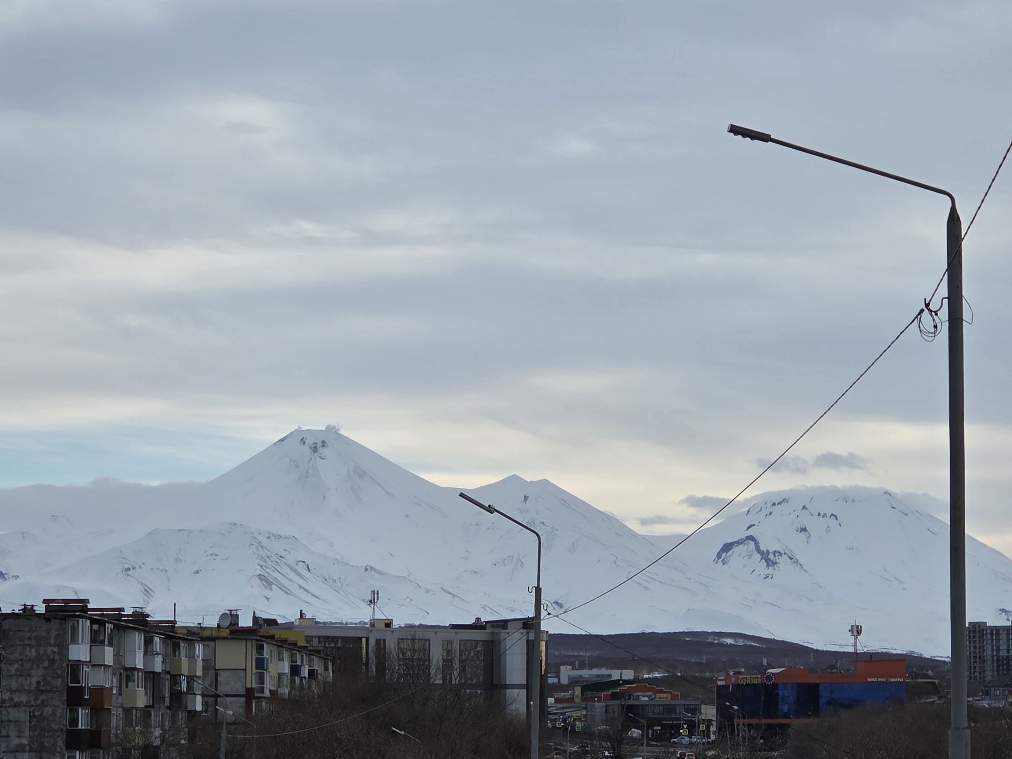 The image captures a city landscape with a backdrop of snow-covered mountains. The sky is overcast, contributing to a muted color palette. In the foreground, apartment buildings with visible wear and tear are present. Street lights are scattered throughout the scene, and vehicles can be observed moving on the roads. Buildings of varying sizes and colors, some with signage, are interspersed within the cityscape. The mountains in the background are the most prominent feature, their peaks covered in snow. The image appears to be taken during the daytime. The location is a city in Unknown, Unknown.